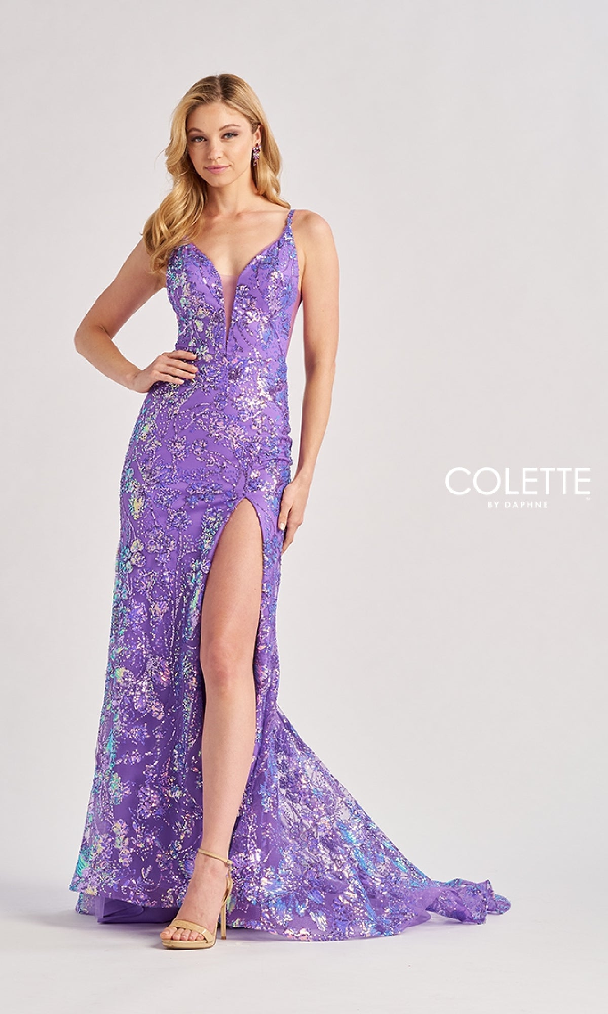 A woman wears the Colette CL8430 long prom dress—a sleeveless, floor-length purple gown with floral sequins and a thigh-high slit—while standing against a plain background.