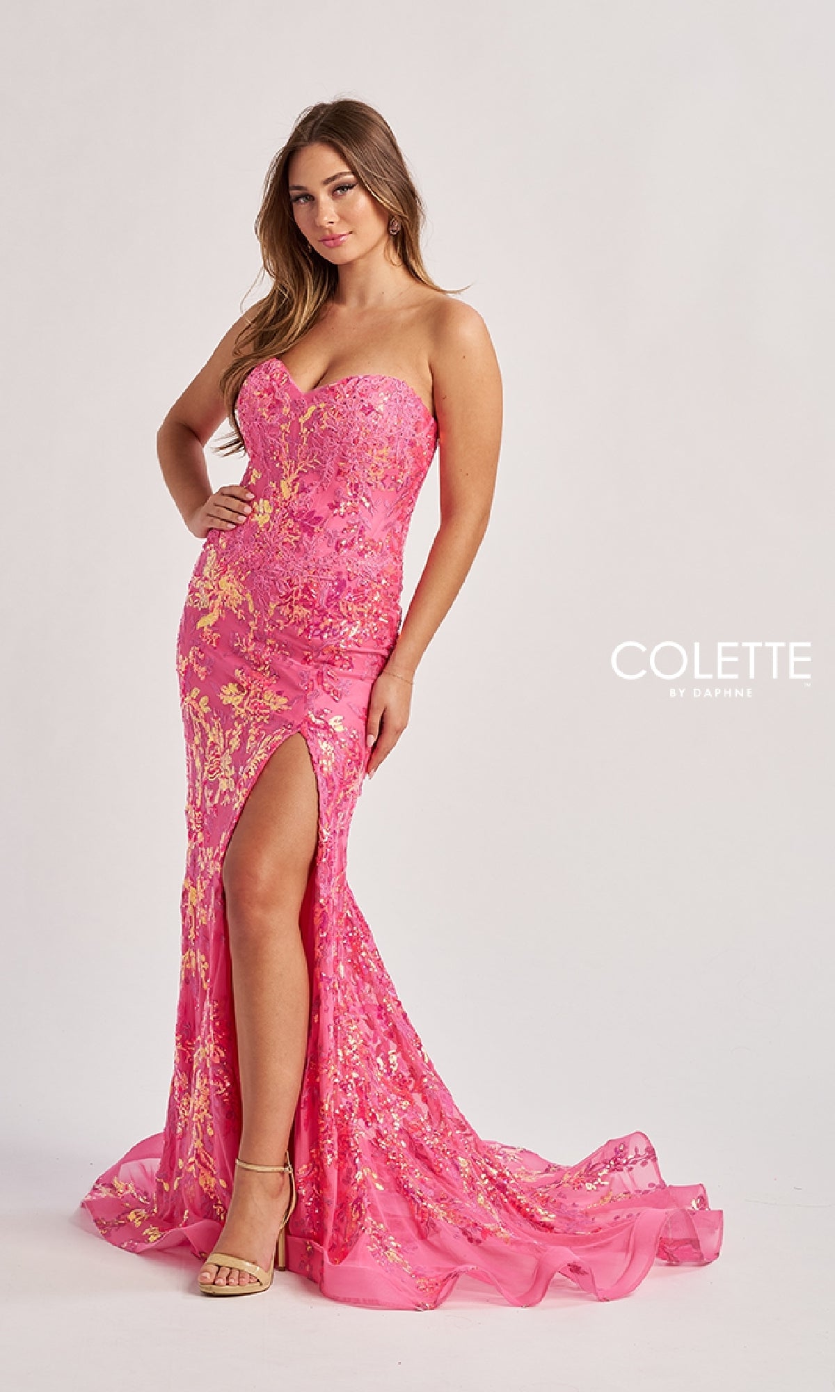 A woman models the Colette CL8440 long prom dress, a strapless mermaid gown with a boned corset bodice, pink fabric adorned with gold floral details, and a thigh-high slit, set against a plain background.