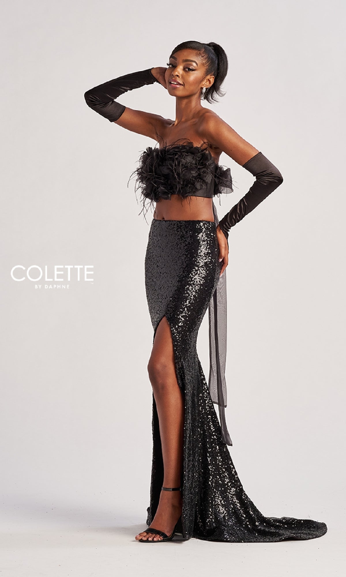 A woman poses in the Colette CL8450 Long Prom Dress, featuring a feathered crop top, long black gloves, and a sequined floor-length skirt with a high slit and train;