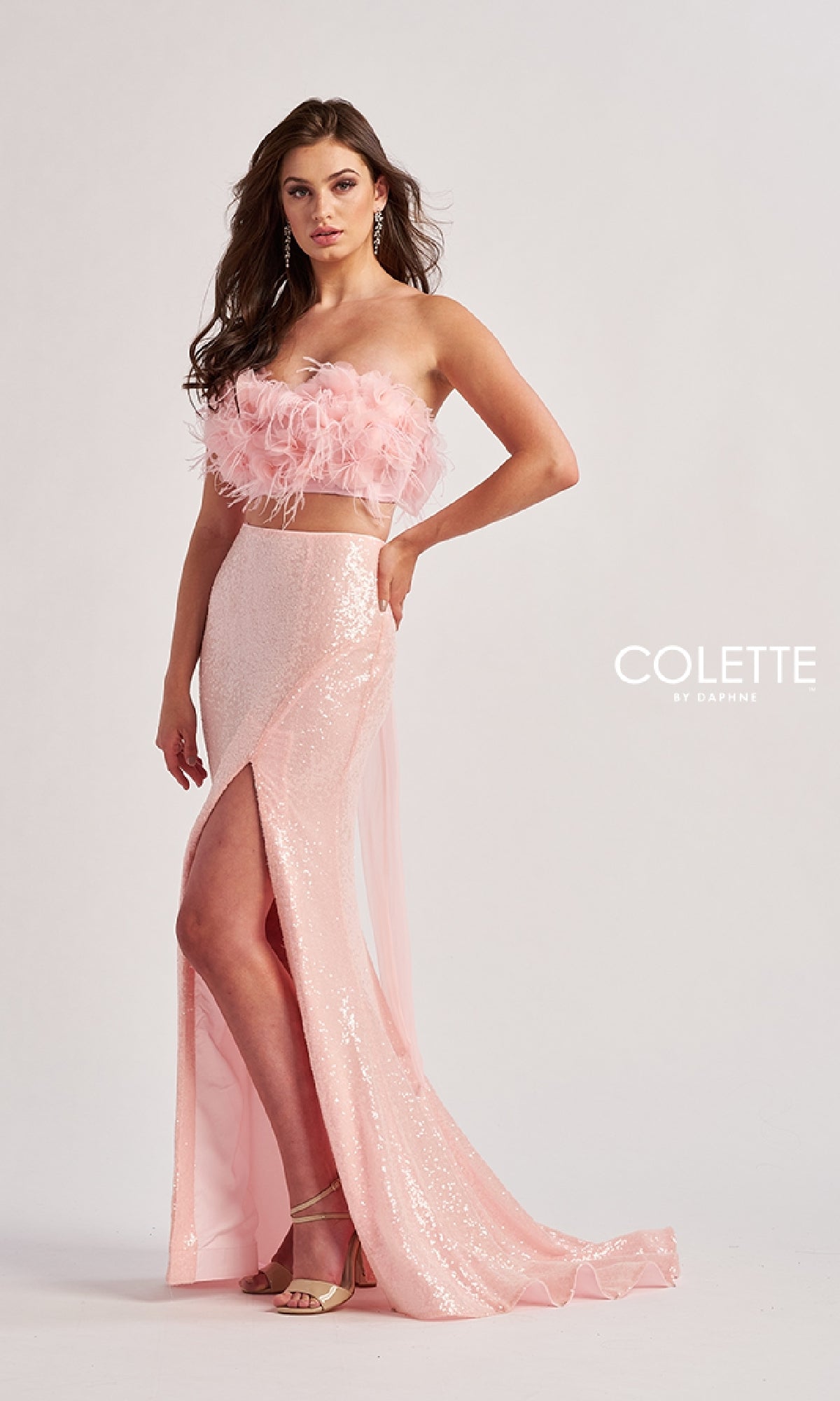 A woman models the Colette CL8450 long prom dress—a pink, strapless two-piece with a feathered crop top and sequined skirt with a thigh-high slit—standing before a plain backdrop,