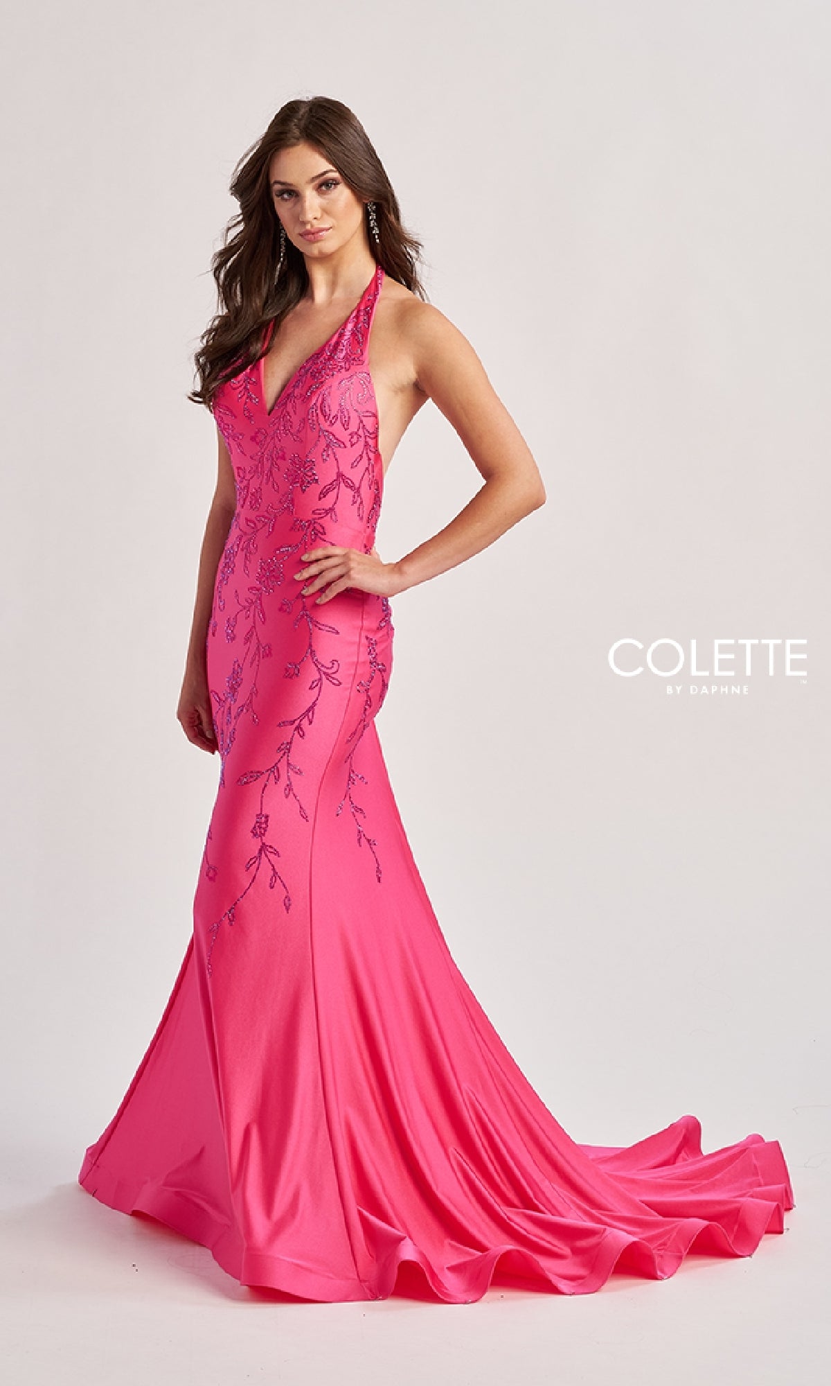 A woman stands against a plain background wearing the Colette CL8455, a floor-length bright pink halter prom dress with embroidery, a v-neckline, fitted bodice, flared skirt, and a small train.