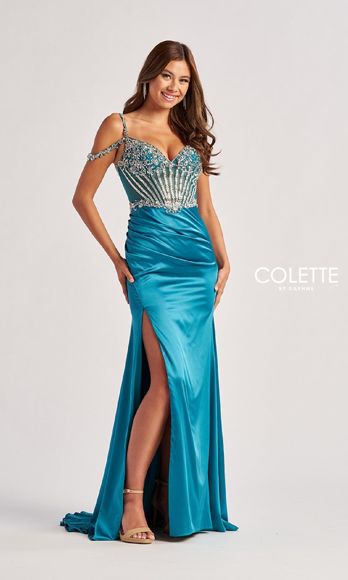A woman poses and smiles in the Colette CL8460 long prom dress featuring a teal color, beaded bodice, lace-up open back, off-shoulder straps, and a thigh-high slit against a plain background.