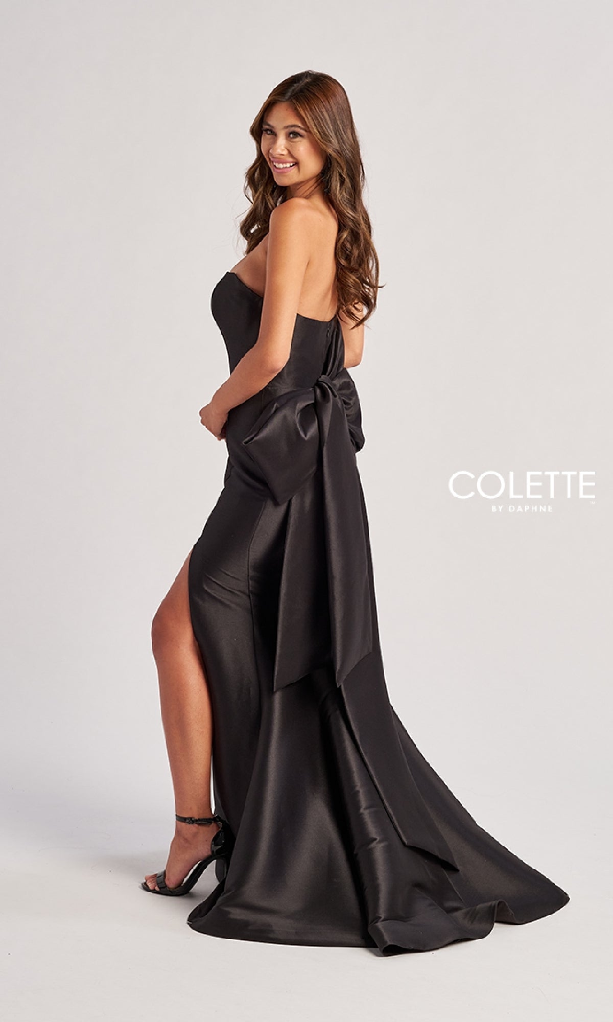 A woman in the Colette CL8470 Back Bow Strapless Long Prom Dress stands sideways and smiles, highlighting the elegant black satin gown with its striking back bow.