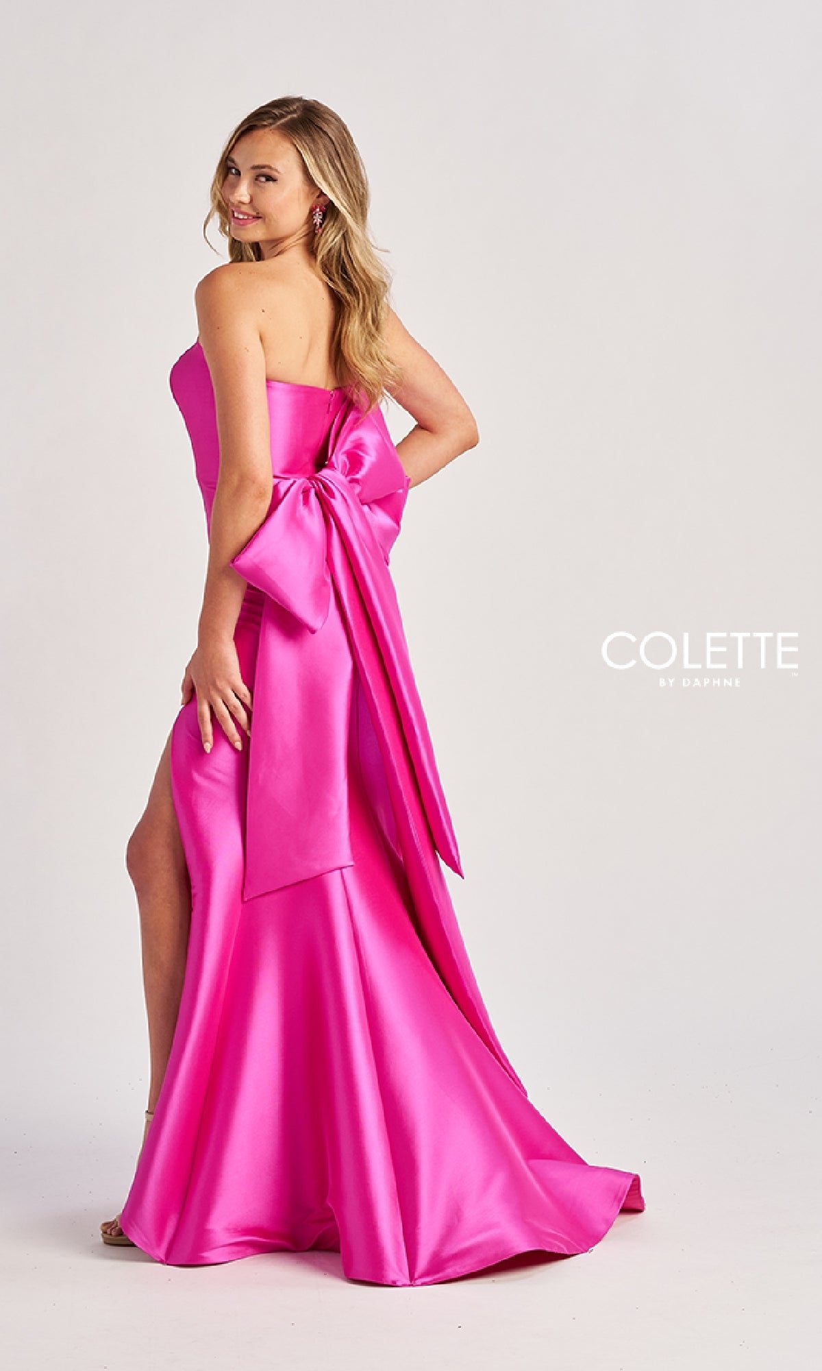 A woman in the Colette CL8470 Back Bow Strapless Long Prom Dress, bright pink with a large back bow, stands sideways and looks over her shoulder. The