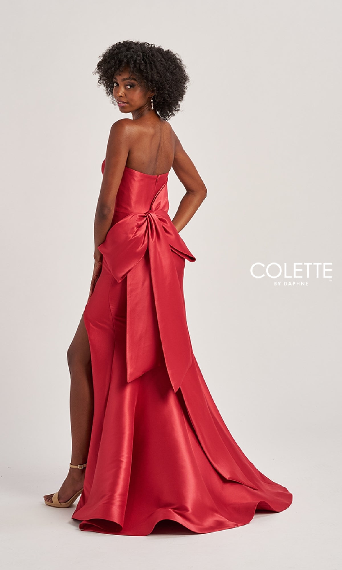 A woman stands against a plain background, looking over her shoulder in the Colette CL8470 Back Bow Strapless Long Prom Dress—an elegant red satin evening gown with a large bow, train, and striking thigh-high slit.