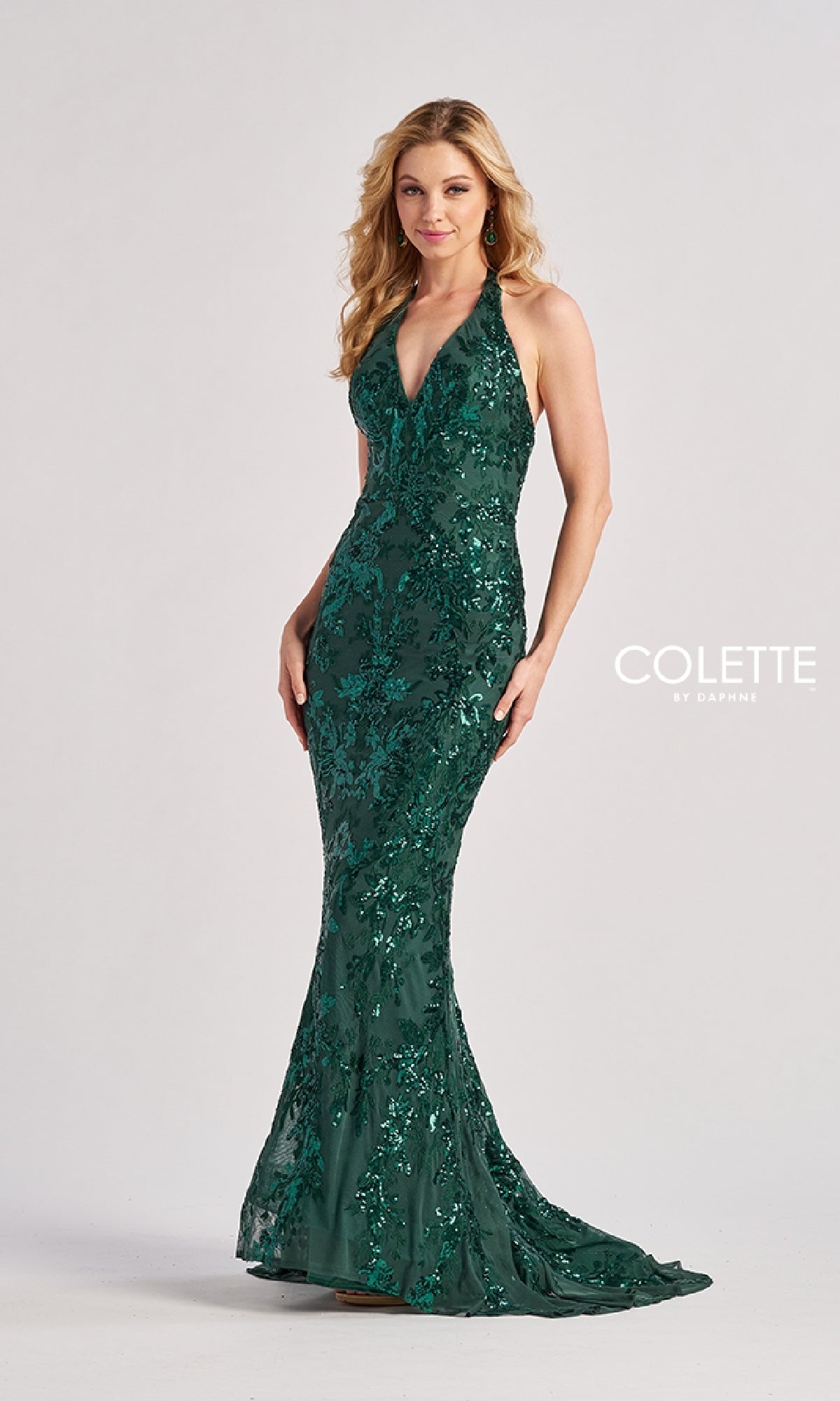 A woman poses against a plain backdrop wearing the Colette CL8495, a floor-length, dark green sequin prom dress with a halter v-neckline and form-fitting silhouette, featuring floral embellishments for added elegance.