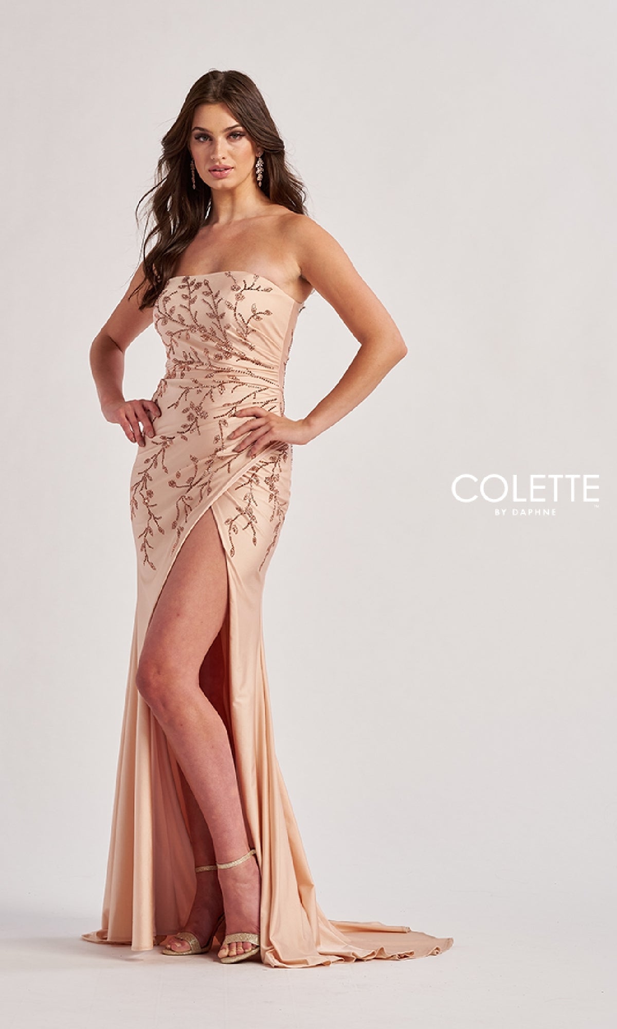 A woman in the Colette CL8500 Long Prom Dress, a strapless blush gown with floral embroidery, sparkling stones, a thigh-high slit, and high heels stands against a plain background.