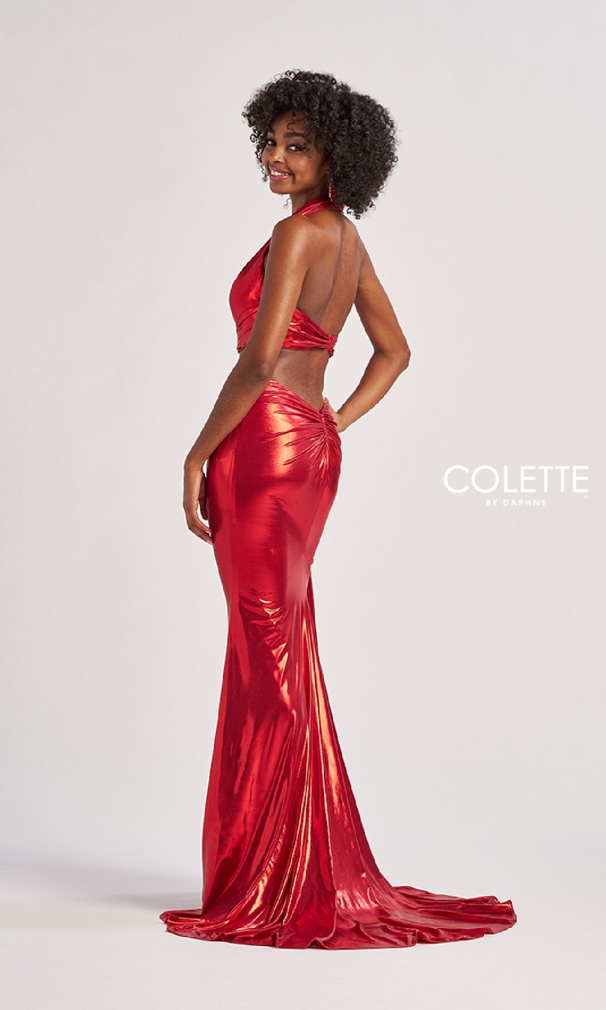 A woman in a shiny red halter prom dress, the Long Prom Dress: Colette CL8505, stands sideways and smiles at the camera.