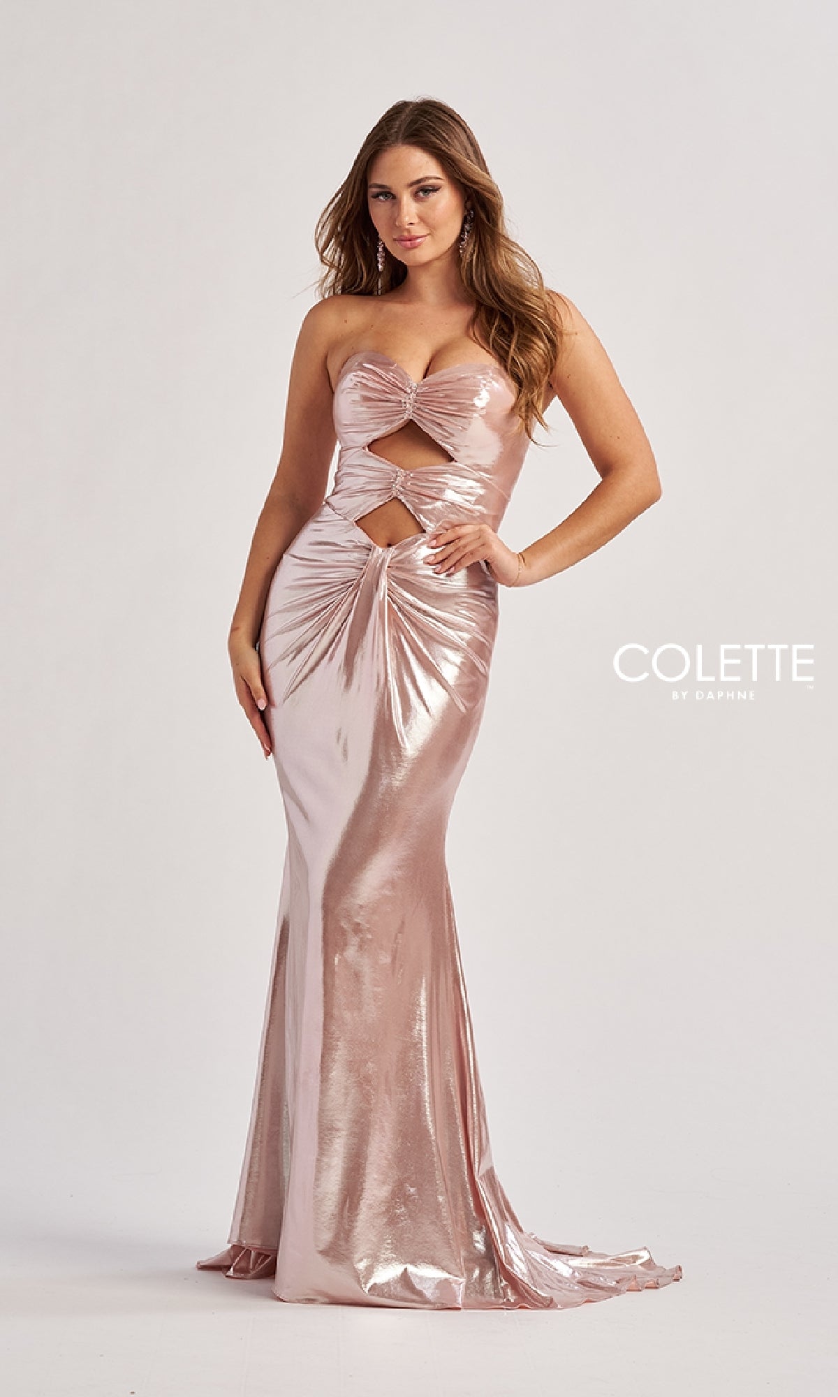 A woman wears the Colette CL8545 long prom dress featuring a shiny light pink metallic finish, strapless design, front cutouts, and ruched details, standing against a plain background.