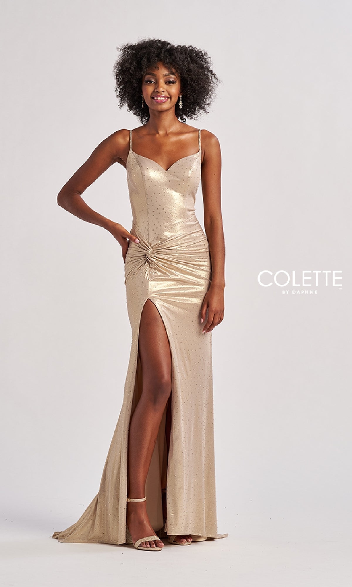 A woman stands against a plain background wearing the Colette CL8575 long prom dress featuring shimmering gold metallic stretch jersey, spaghetti straps, a ruched waist, and a thigh-high side slit.