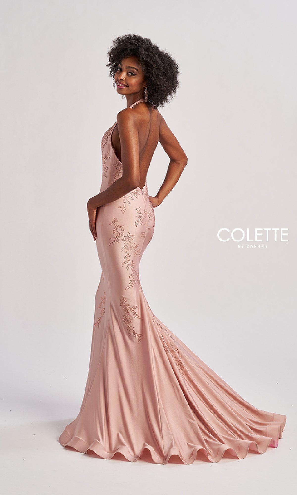 A woman models the Colette CL8580 long prom dress, a light pink, backless, floor-length mermaid gown with floral embroidery and beaded sequin appliques. She stands sideways, looking over her shoulder against a plain background.