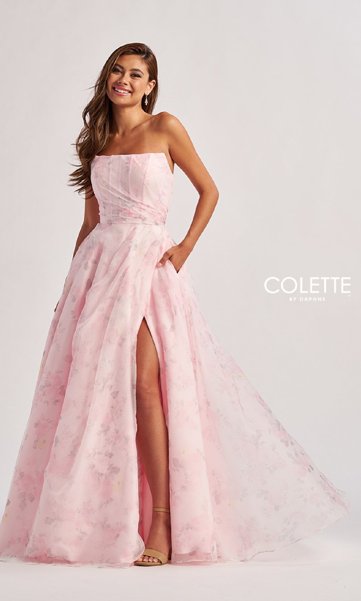Woman wearing the Colette CL8635 long prom dress—a strapless, light pink floral a-line gown with pockets and a thigh-high slit—stands against a plain background.