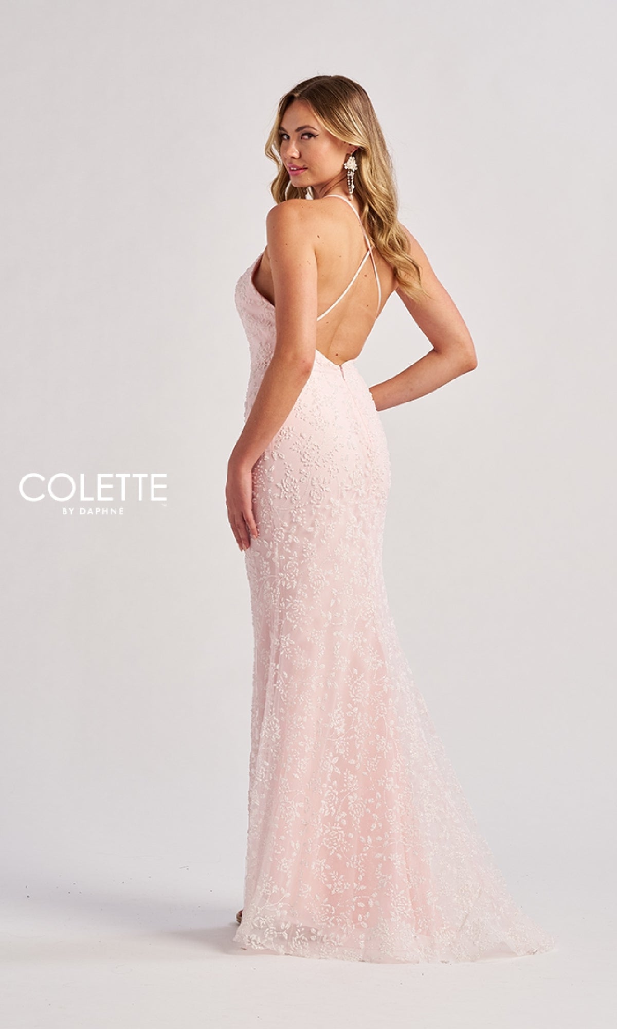 A woman stands in a studio wearing the Colette CL8655, a light pink, floor-length lace prom dress with a v-neckline and thin crisscross back straps, set against a plain background.
