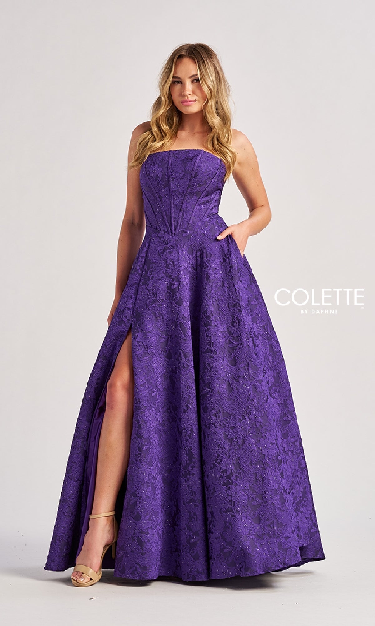 A woman models the Colette CL8675 long prom dress, featuring a purple strapless bodice, textured pattern, floor-length a-line skirt with high slit, against a plain background.
