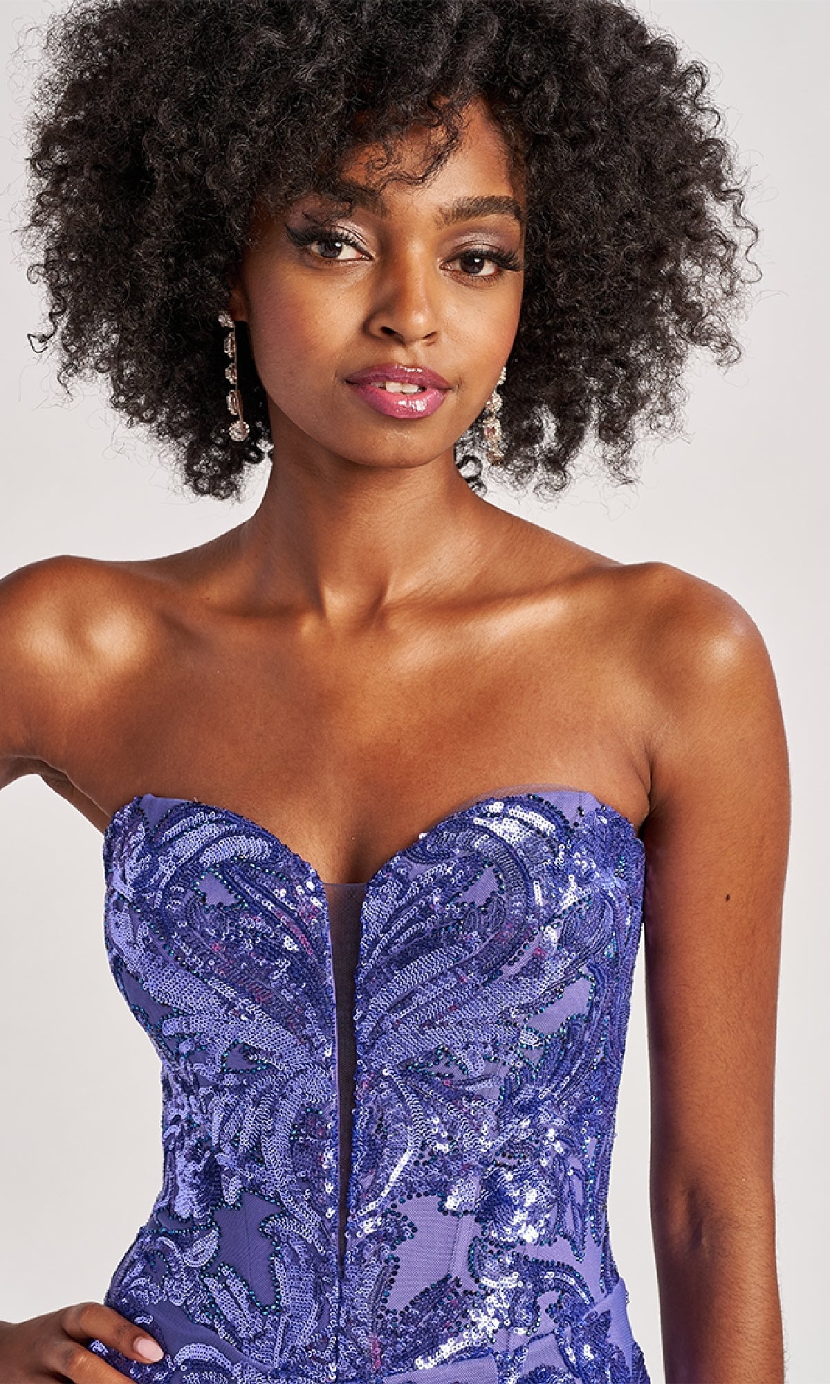 A woman with curly hair wears the Colette CL8680 long prom dress, a strapless purple sequin gown, paired with dangling earrings against a neutral background.