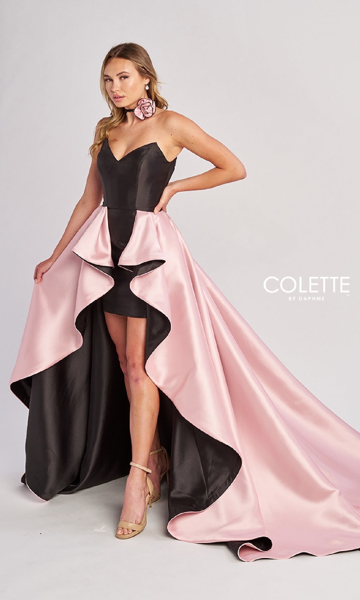 Wearing the Colette CL8695 long prom dress featuring a dramatic pink satin overskirt, a woman pairs it with ankle-strap heels and floral earrings, posing against a plain light background.