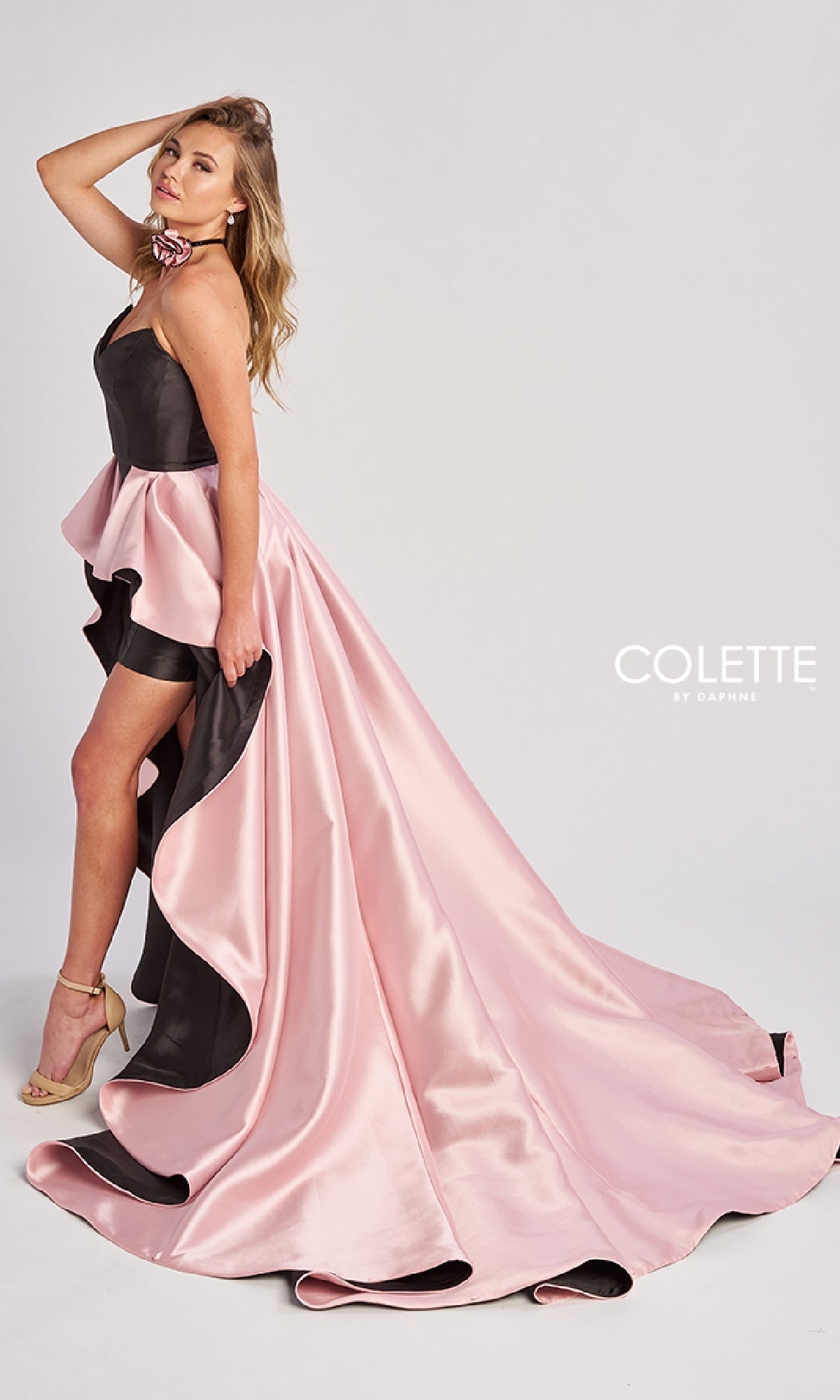 A woman poses in the Long Prom Dress: Colette CL8695, which features a strapless design, high-low hem with a flowing overskirt. She completes her look with gold heels and holds her hand near her face.