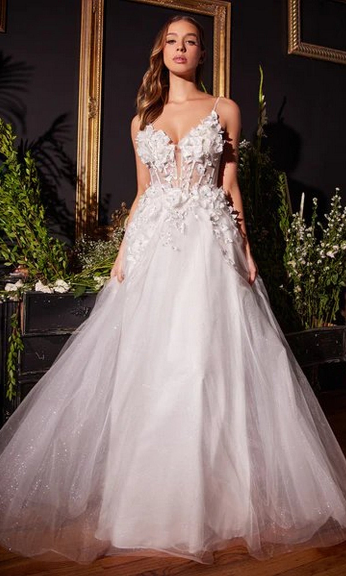 A woman stands indoors among lush greenery and empty picture frames, wearing the Off-White Long Floral Prom Ball Gown CM321W with a floral-embellished skirt.