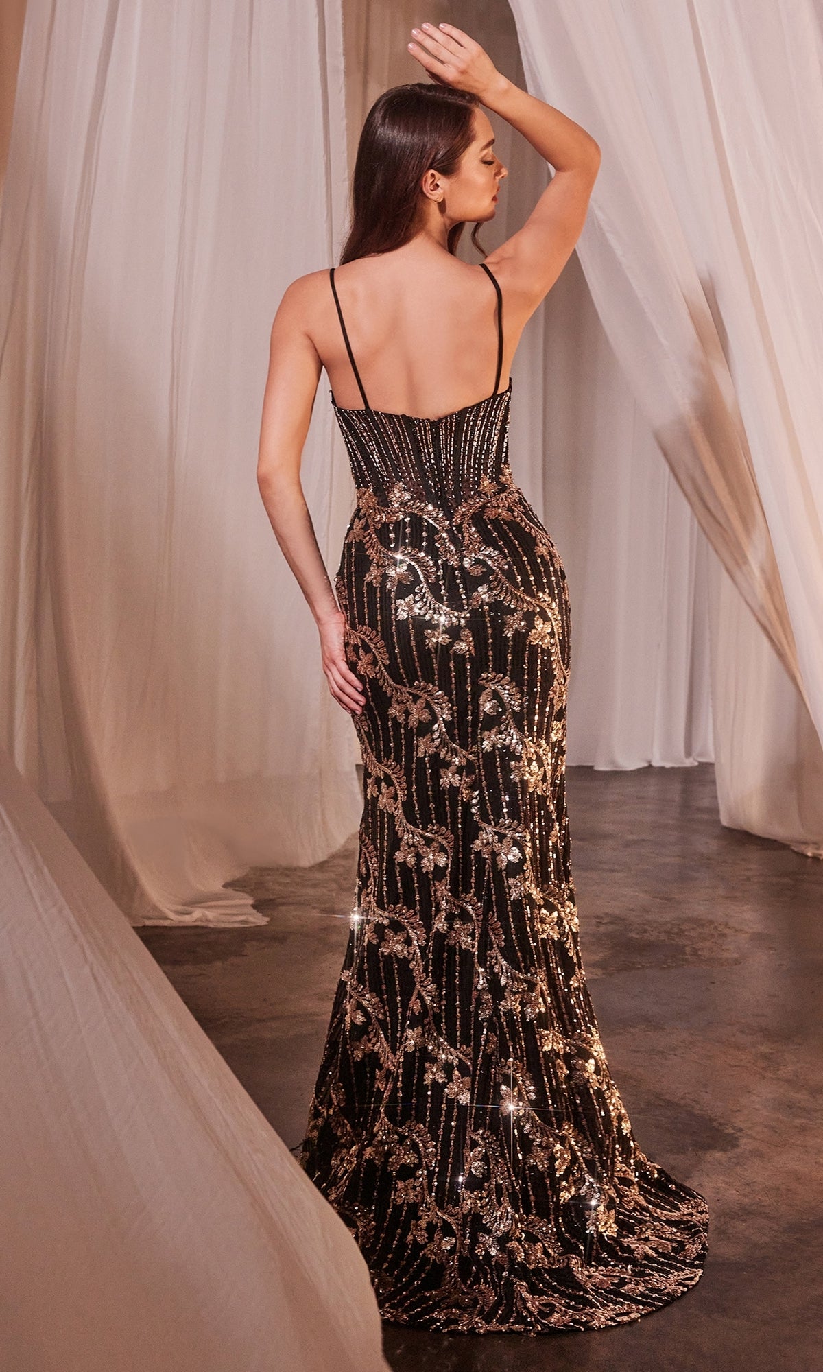 A woman stands with her back to the camera, wearing the Ladivine CM358 Gold-Trimmed Long Black Prom Dress with metallic sequin embellishments and thin straps, surrounded by draped white curtains.