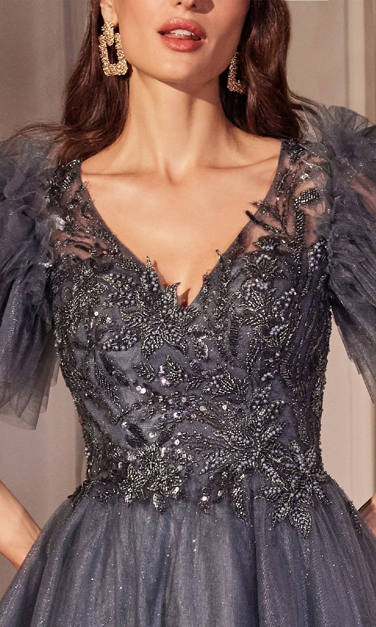 A woman wears the Ladivine CM359 Flutter-Sleeve Blue Prom Ball Gown, a dark sheer dress with flutter tulle sleeves, a sequin-lace bodice, and intricate floral embellishments, paired with large gold earrings.