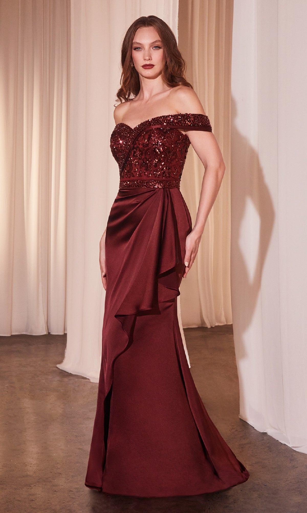 A woman stands before white curtains wearing the Off-Shoulder Draped Long Prom Gown Ladivine CR877 in deep red, featuring a beaded sequin bodice and a flowing skirt.