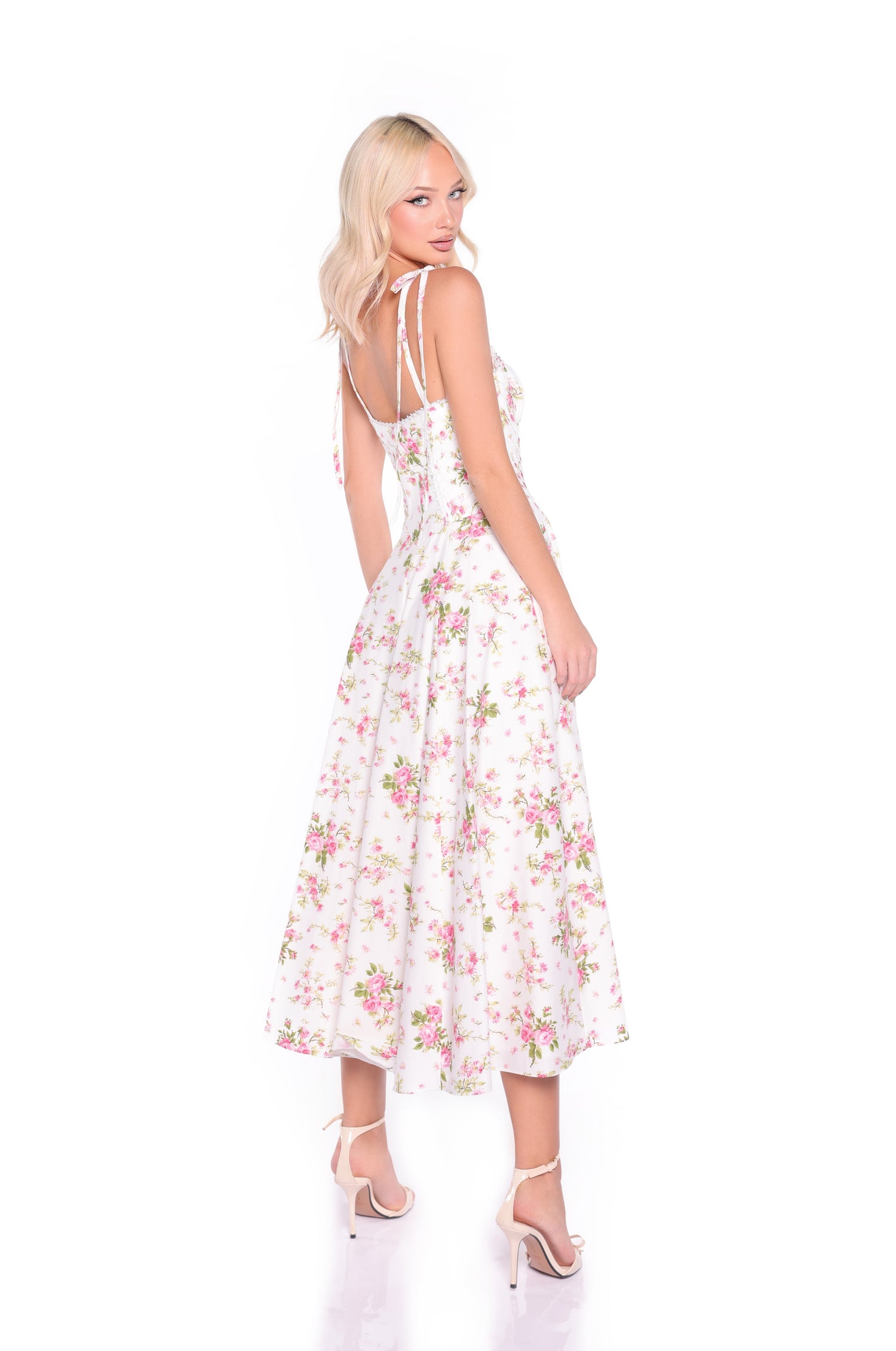 Woman wears the Ivory Blush Floral Midi Casual Dress: Cecilia, featuring tie straps and a corset-style bodice, looking over her shoulder in heels against a white background. Ideal as a summer midi dress for any occasion.