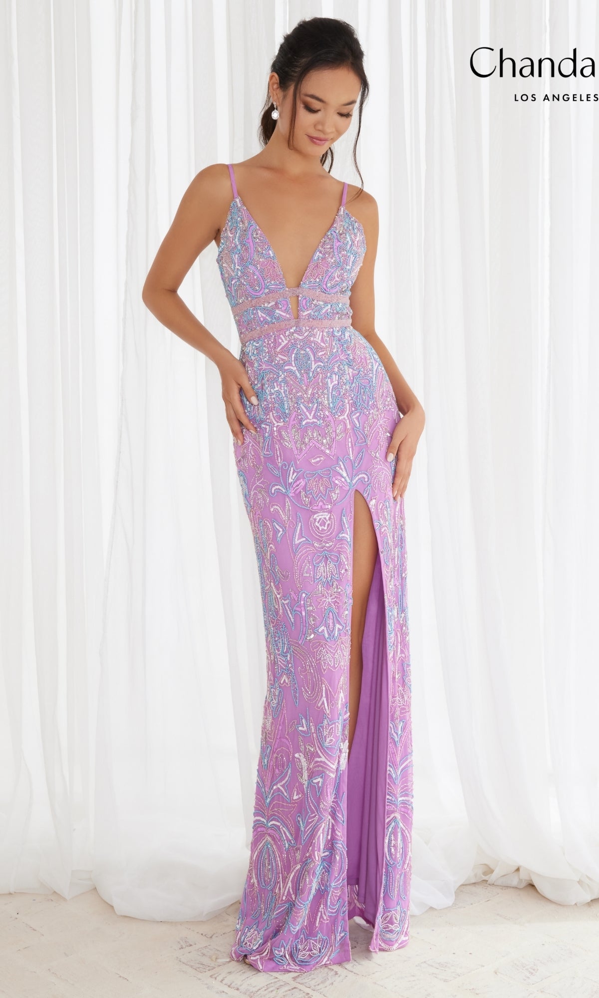 A woman poses in front of sheer white curtains wearing the Light Purple Long Beaded Dress 30173 by Chandalier, featuring a sleeveless design, deep V-neckline, high slit, and intricate beadwork.