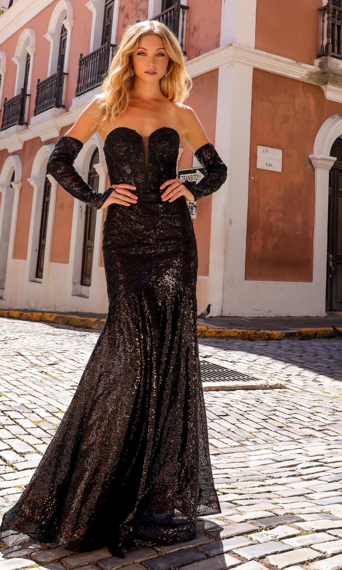 A woman in the Nox Anabel Strapless Sequin Lace Prom Dress D1263 with matching gloves stands on a cobblestone street by a peach-colored building.