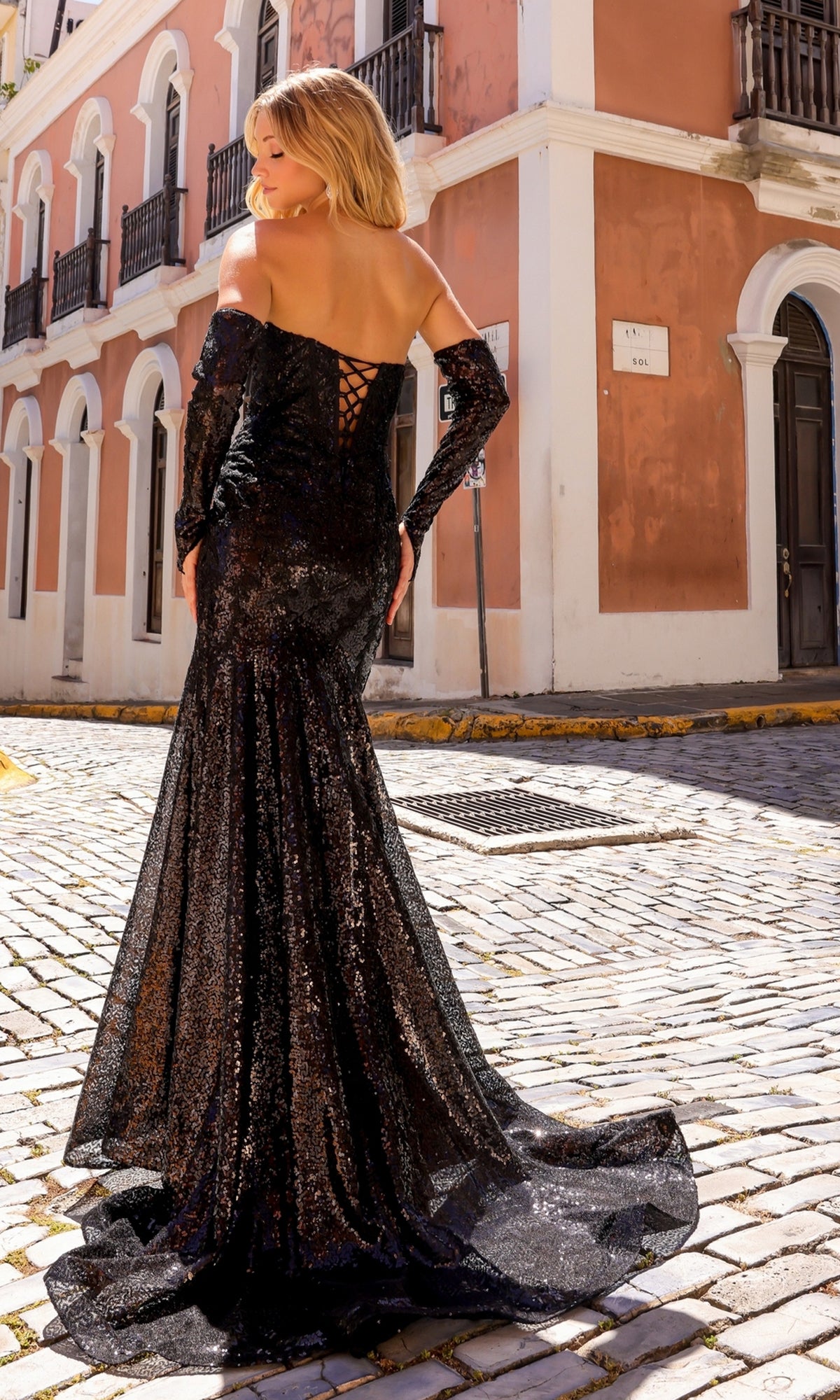 A person in a black Nox Anabel Strapless Sequin Lace Prom Dress D1263 stands on a cobblestone street before a peach-colored building with white trim.