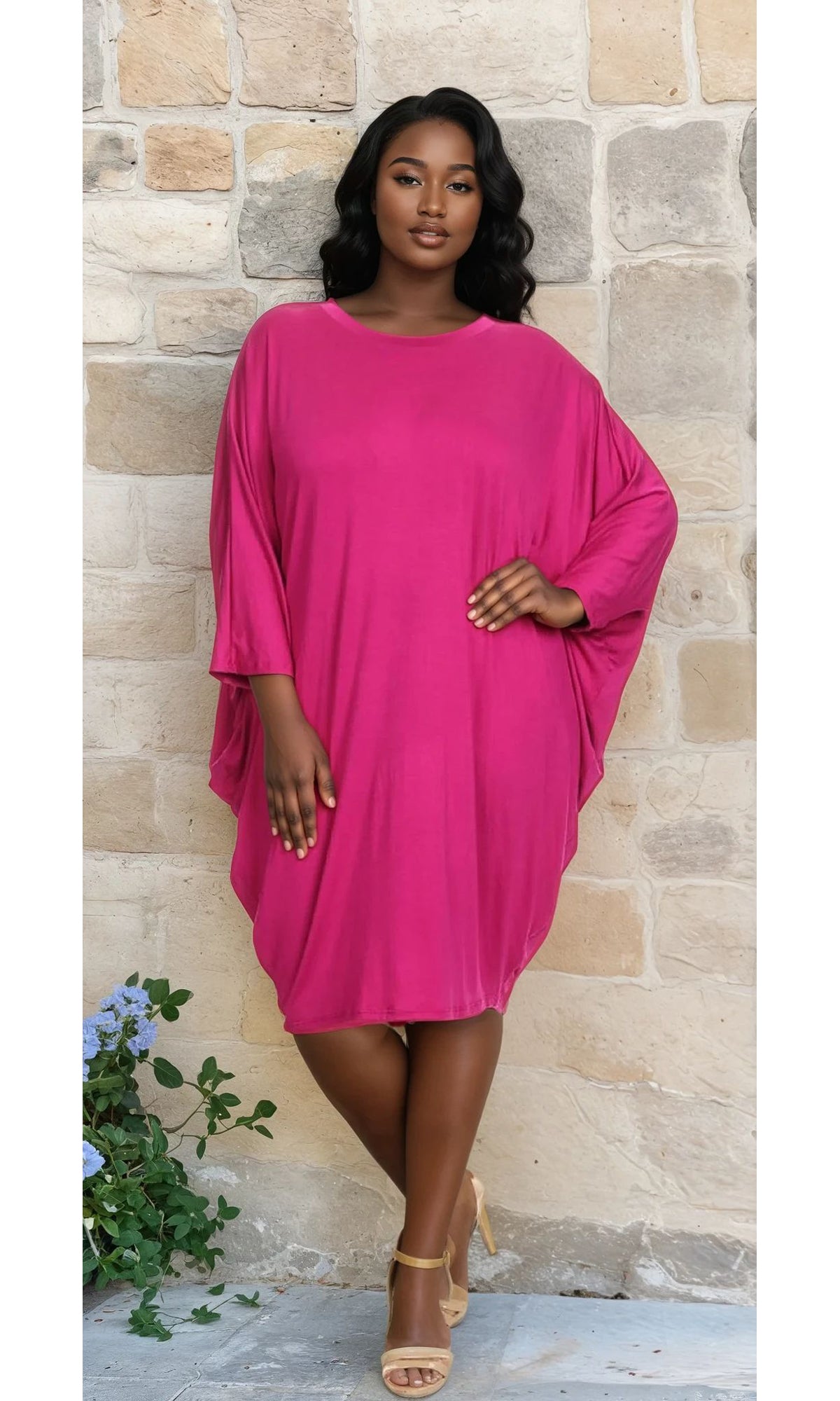 A woman poses by a stone wall, hand on her hip, wearing the Short Casual Plus Size Dress with Dolman Sleeves in bright pink and beige sandals.