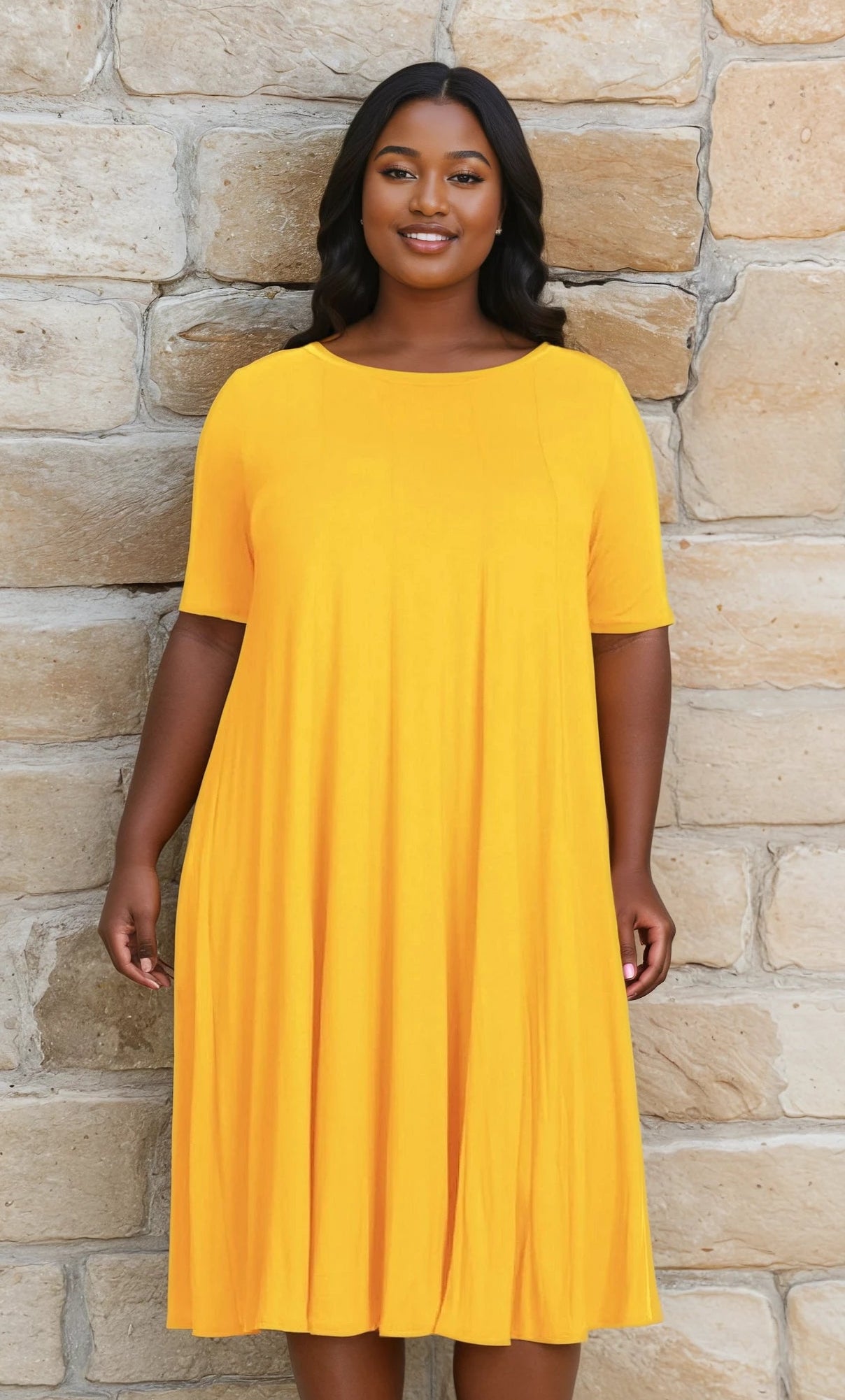 A woman stands in front of a stone wall, smiling at the camera in a Plus-Size Jersey Knit Oversized A-Line Casual Dress, featuring short sleeves and a knee-length bright yellow design.
