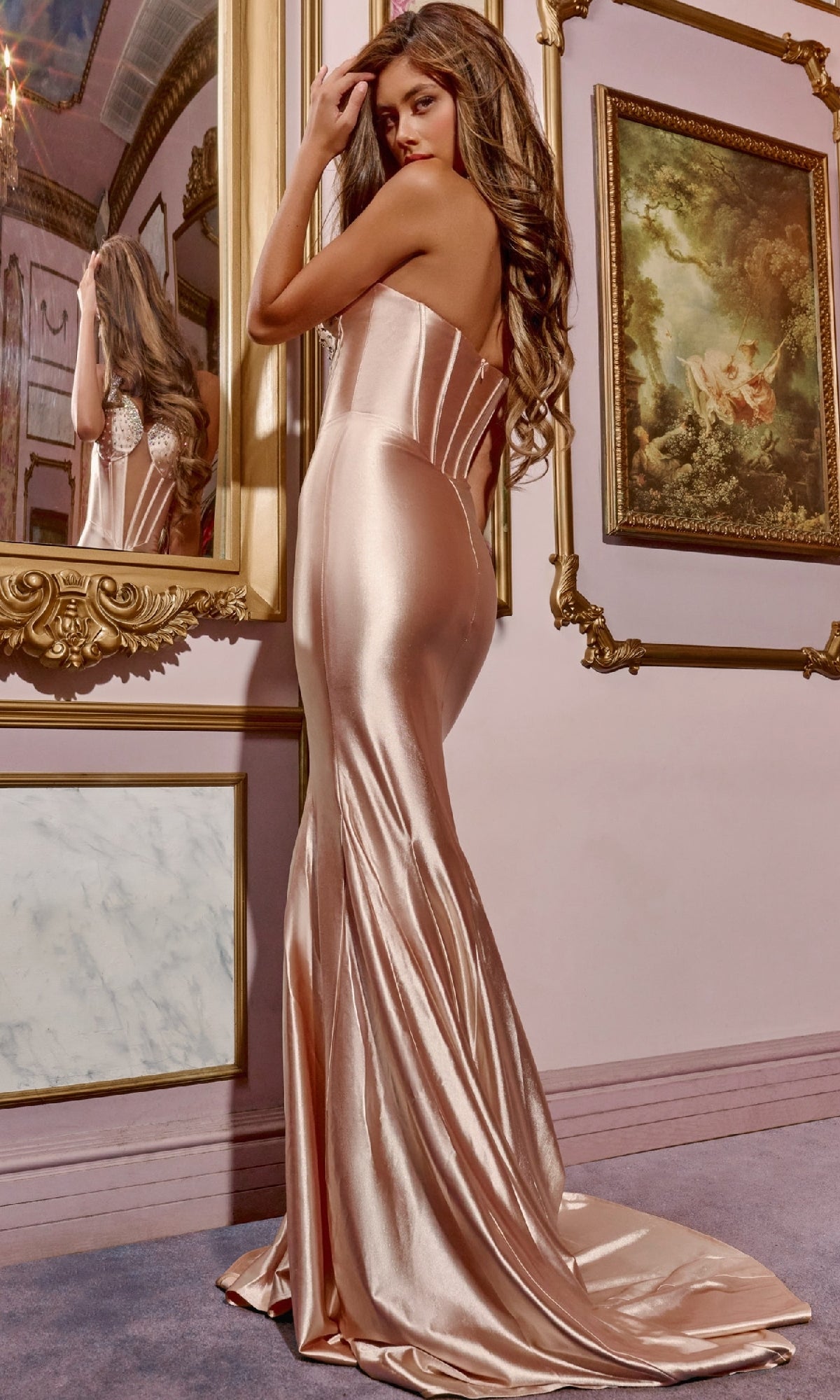 Wearing the Long Prom Dress: Jovani D5018, a woman in a shiny, strapless gown with a sleek corset bodice stands before ornate framed art, glancing over her shoulder.