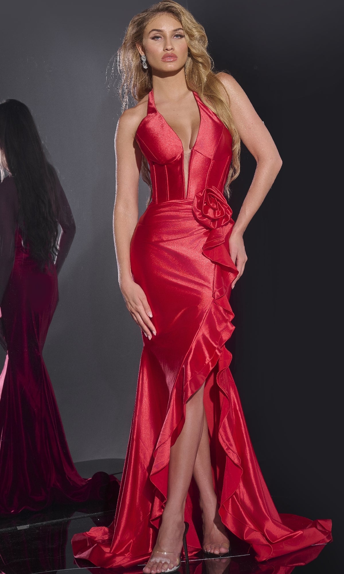 A woman stands against a dark background wearing the Jovani D5306 long prom dress—form-fitting in deep red with a corset bodice, plunging neckline, ruffle accents, and a high slit.