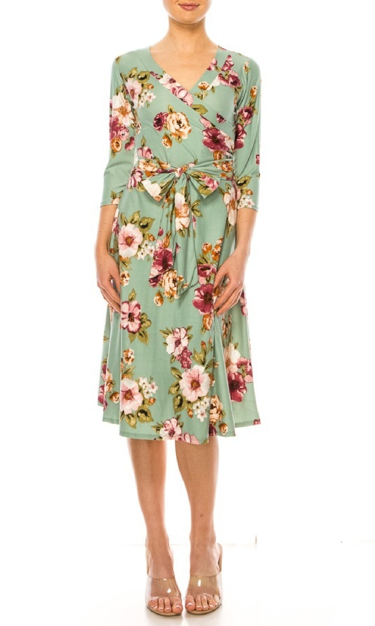 A woman models the Sage Green Floral Print Faux-Wrap Casual Dress with three-quarter sleeves, a waist tie, and v-neckline, paired with nude open-toe heels against a white background.