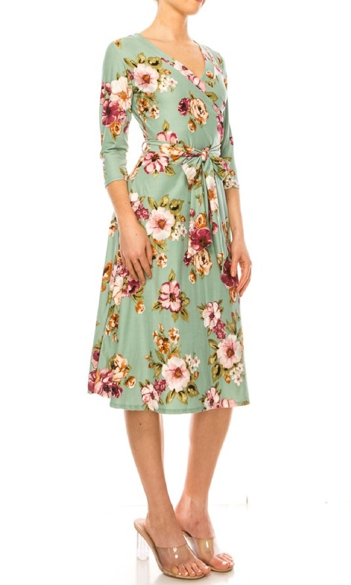 A woman wearing the Sage Green Floral Print Faux-Wrap Casual Dress with three-quarter sleeves and a waist tie, styled with clear heeled sandals.