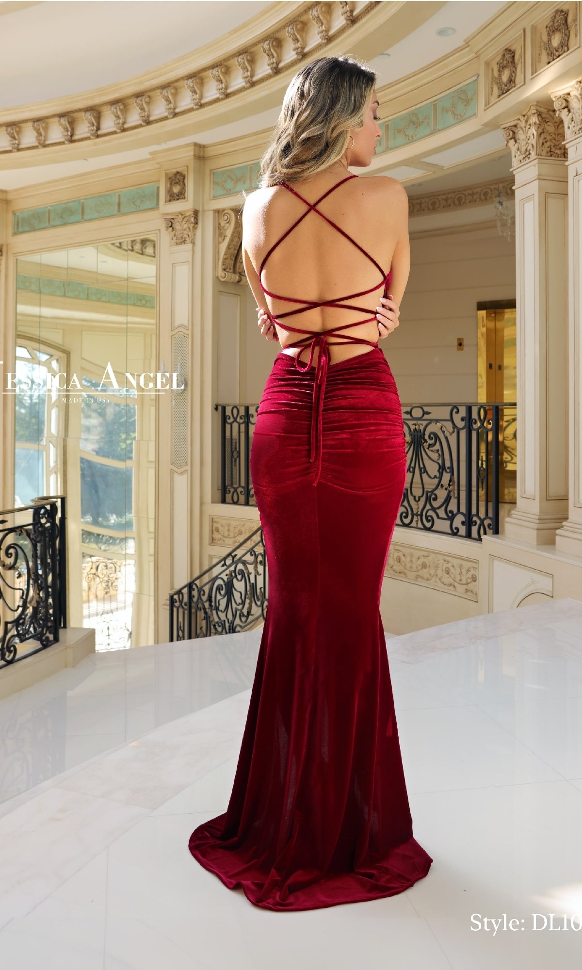 A woman indoors faces away from the camera, wearing the Jessica Angel DL108 Tight Long Velvet Prom Dress—a floor-length, backless red gown with crisscross straps and ruched detailing.