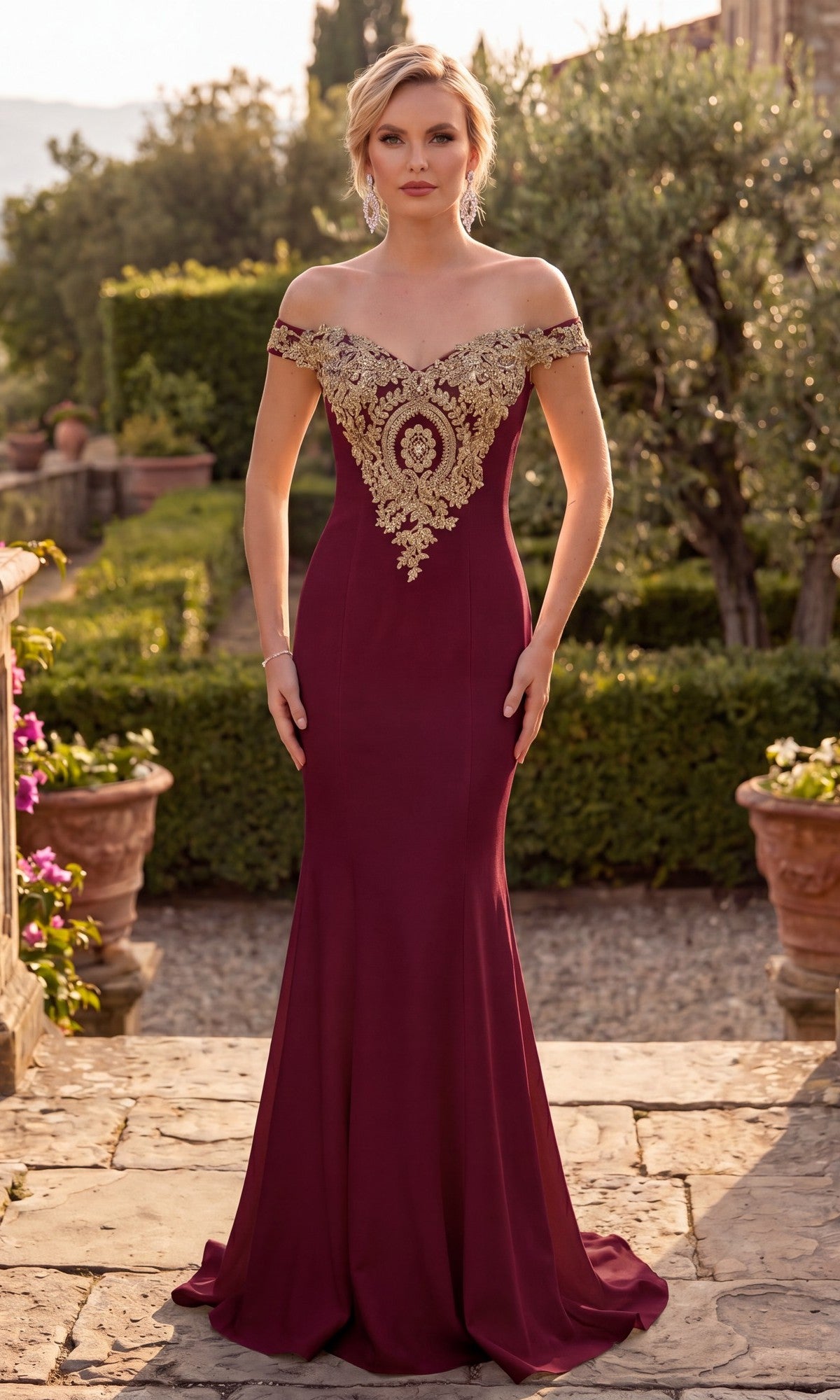 A woman stands outdoors on a stone path, surrounded by greenery and potted plants, wearing the Chicas DM4011 long formal dress in burgundy, featuring an off-the-shoulder design with an embroidered bodice and gold appliques.