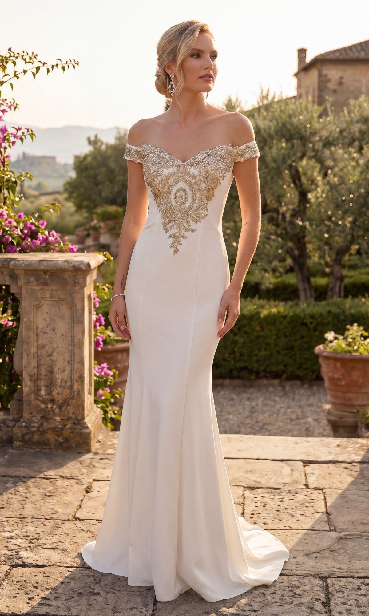 A woman in the Long Formal Dress: Chicas DM4011, a stunning white off-the-shoulder gown featuring a beautifully embroidered bodice and shimmering gold appliques, stands outdoors on a stone patio surrounded by lush greenery and blooming flowers.