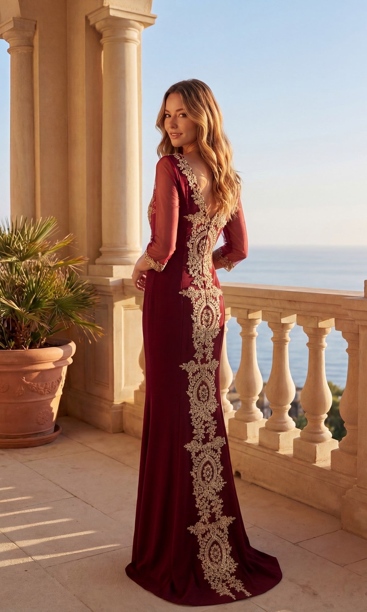 A woman in the Long Formal Dress: Chicas DM4020—burgundy with gold embroidery and a v-neckline—stands on a stone balcony overlooking the ocean, with a potted plant nearby.