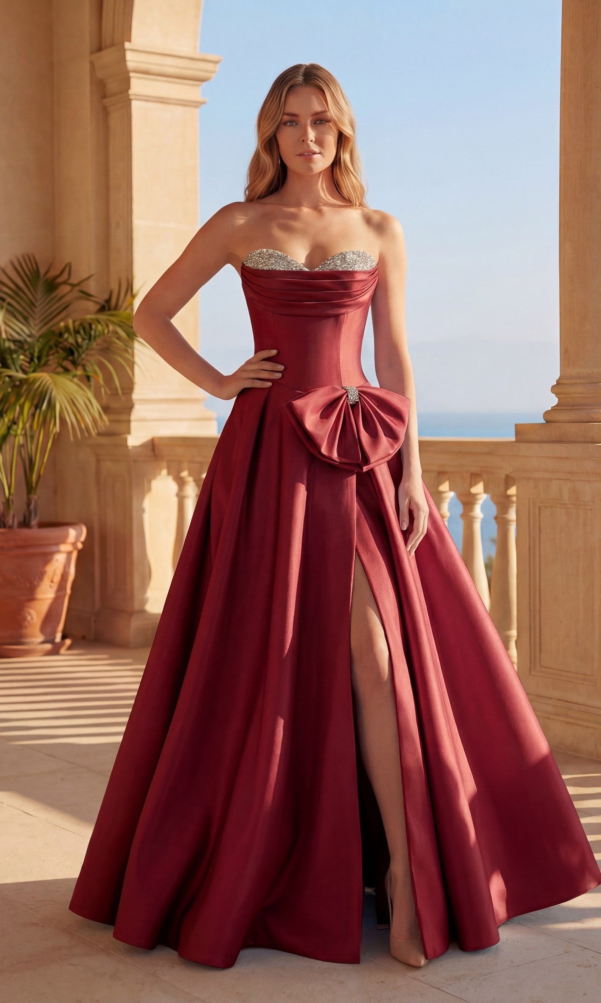 A woman stands on a terrace wearing the Long Prom Dress: Chicas DM9003 in burgundy, a strapless mikado ball gown with a sweetheart neckline, thigh-high slit, and silver beaded bodice detail.