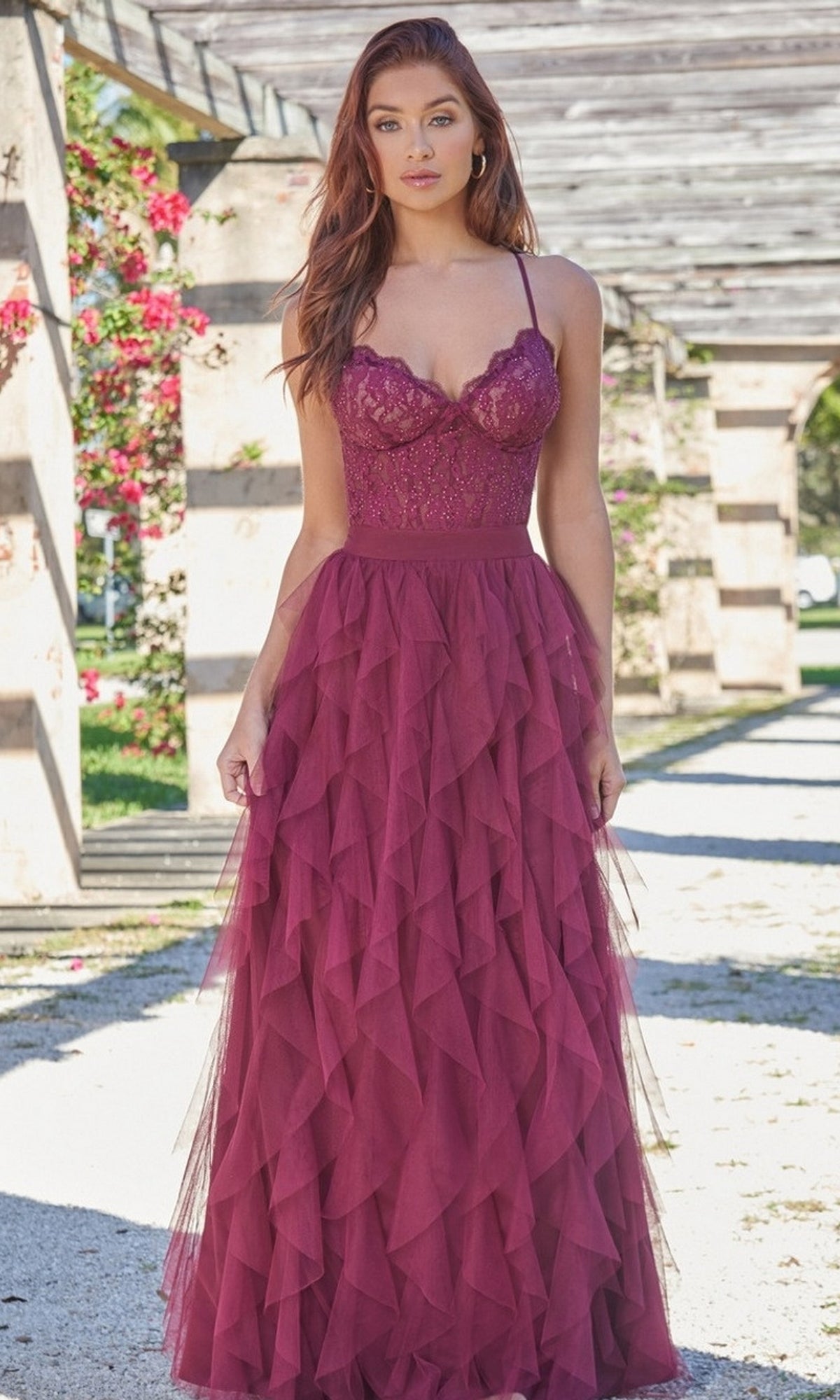 A woman stands outdoors wearing the Velvi Lace-Bodice Long Ruffled Prom Dress Dahlia, featuring a mulberry purple A-line silhouette with a lace bodice and layered tulle skirt.