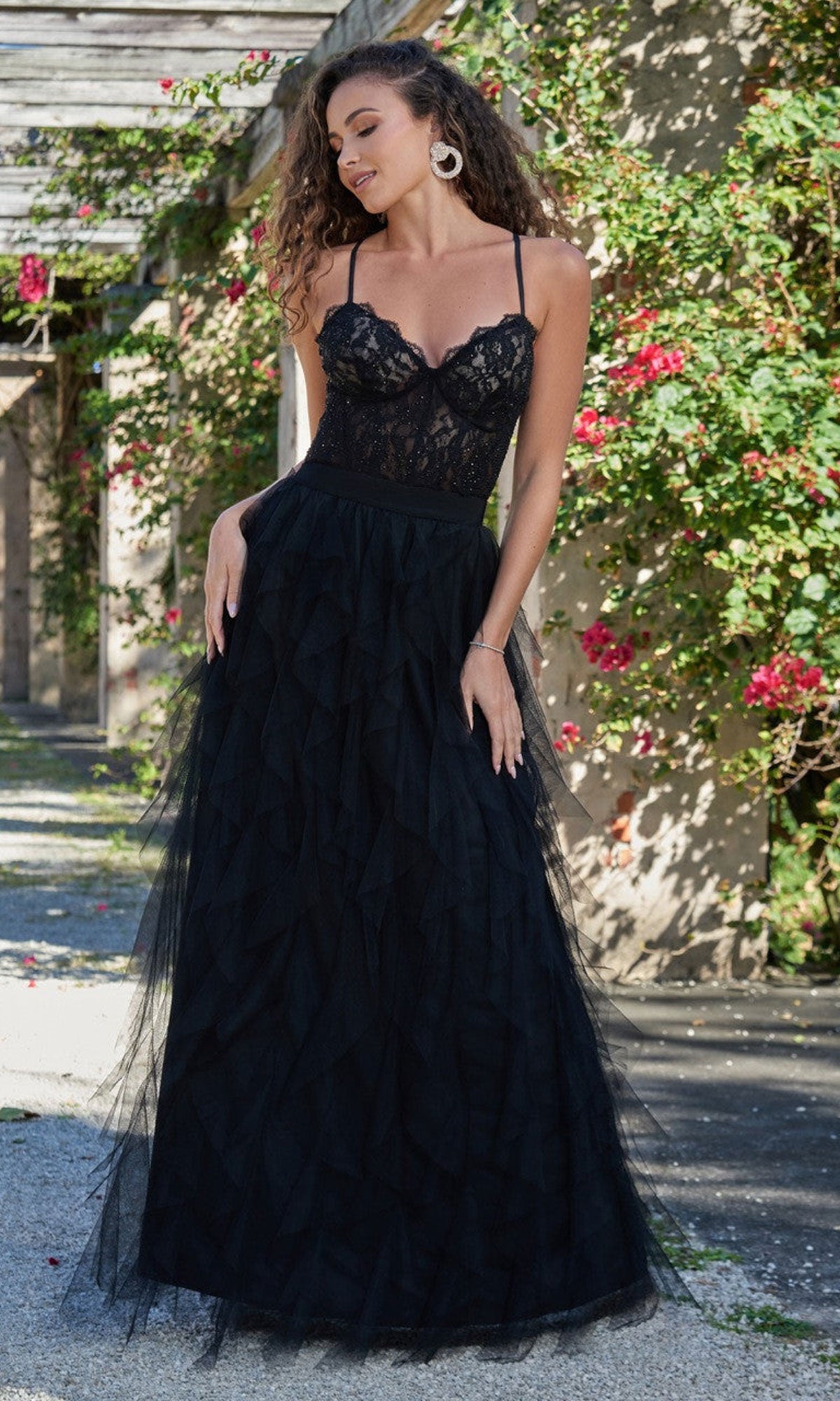 A woman stands outdoors in the Velvi Lace-Bodice Long Ruffled Prom Dress Dahlia, featuring a black lace bodice and tulle skirt, surrounded by greenery and flowers.