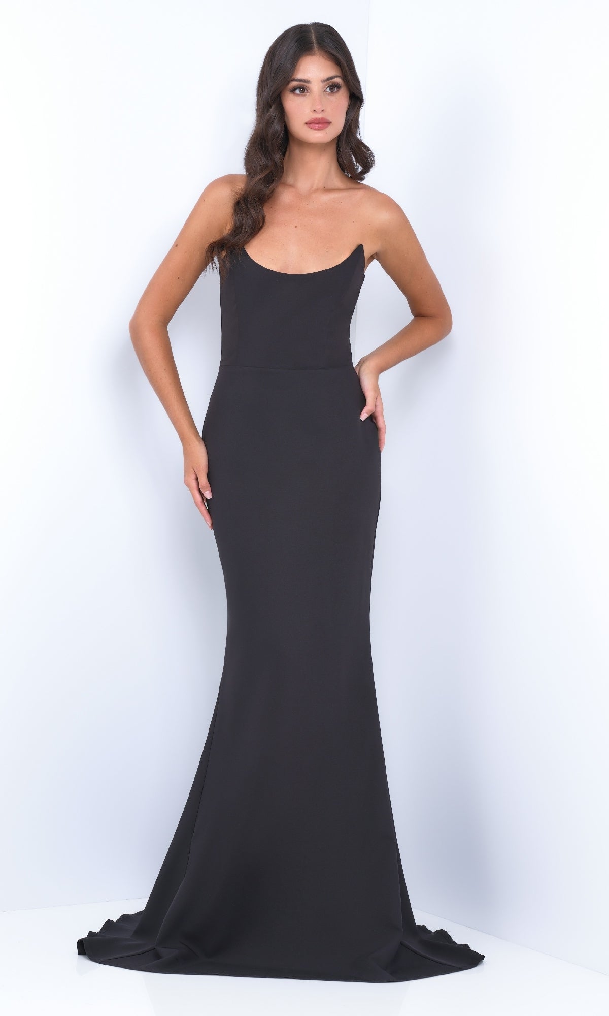A woman with long wavy hair models the Strapless Classic Black Prom Dress: Daris by Velvi, a fitted, floor-length gown, standing against a plain white background.