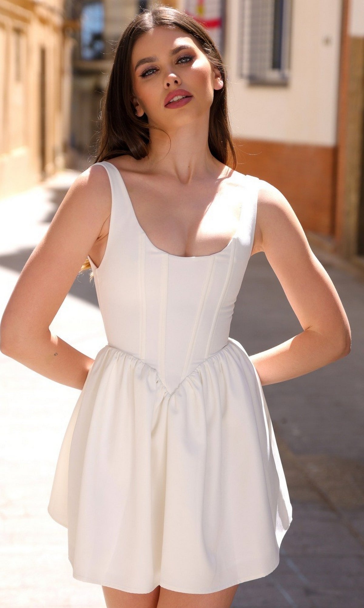A woman stands outdoors on a sunlit street, wearing the Velvi A-Line Mini Graduation Party Dress Darling—a sleeveless, white fitted cocktail dress with a flared skirt, ideal as a short semi-formal dress for special occasions.