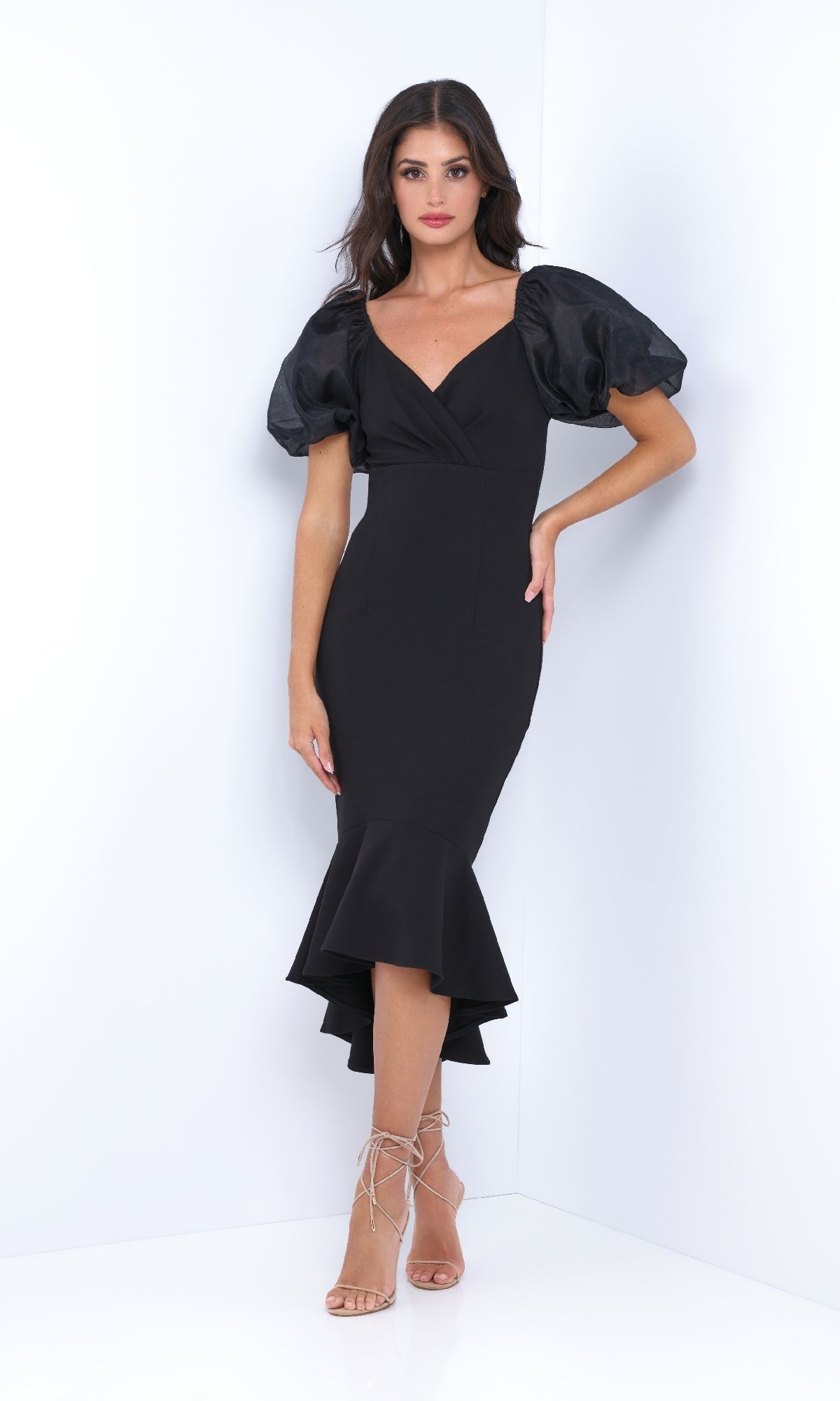 A woman wears the Velvi Short Sleeve Midi Semi-Formal Dress Diana, featuring a fitted black design with puff sleeves, V-neckline, and ruffled hem, styled with strappy high-heeled sandals against a plain white background.