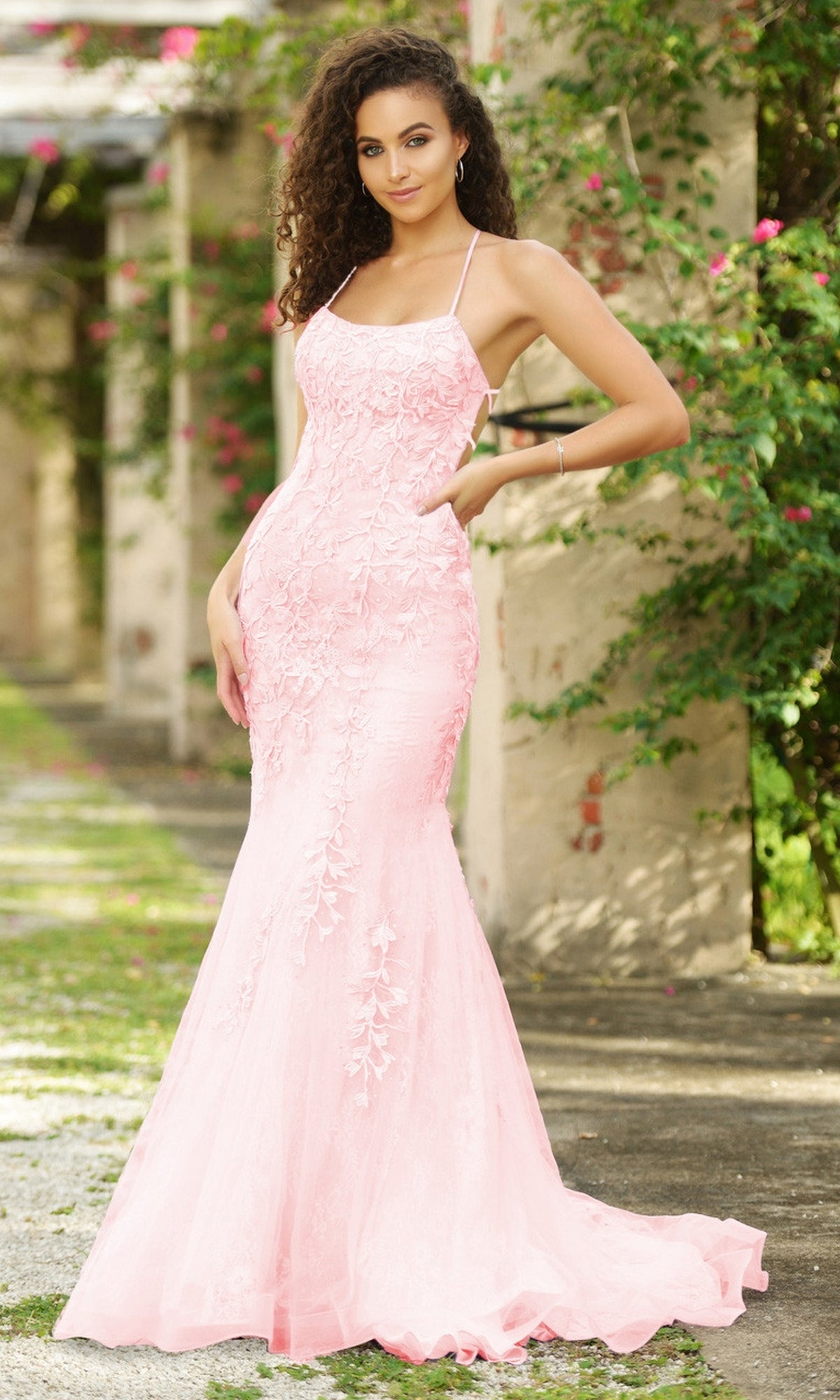 A woman wearing the Beaded Lace Long Prom Dress Dutchess, a light pink fitted gown with floral lace details, stands outdoors before stone columns and lush greenery.