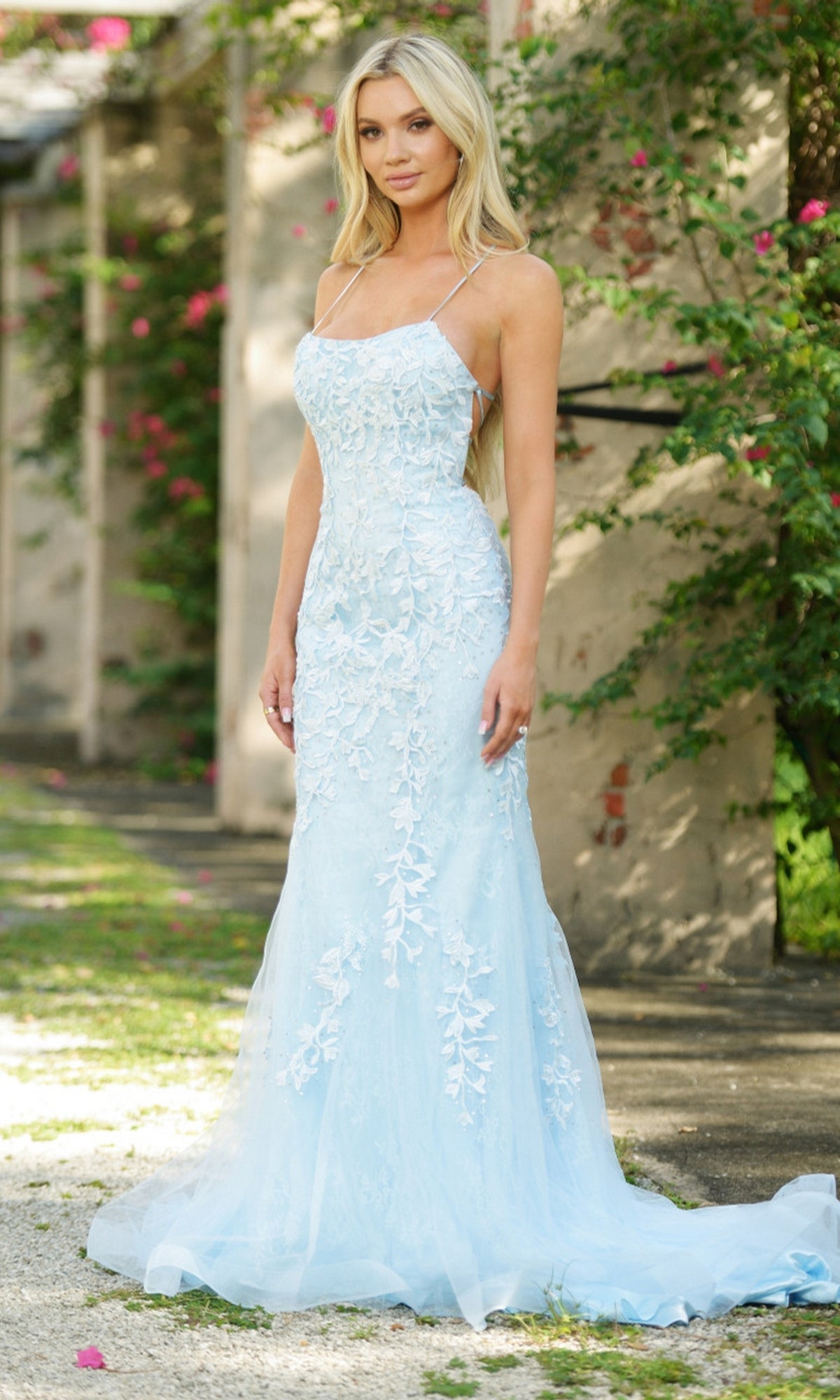 A woman outdoors wears the Beaded Lace Long Prom Dress Dutchess—a light blue, floor-length gown with thin straps and a mermaid silhouette—set against greenery and a stone wall.