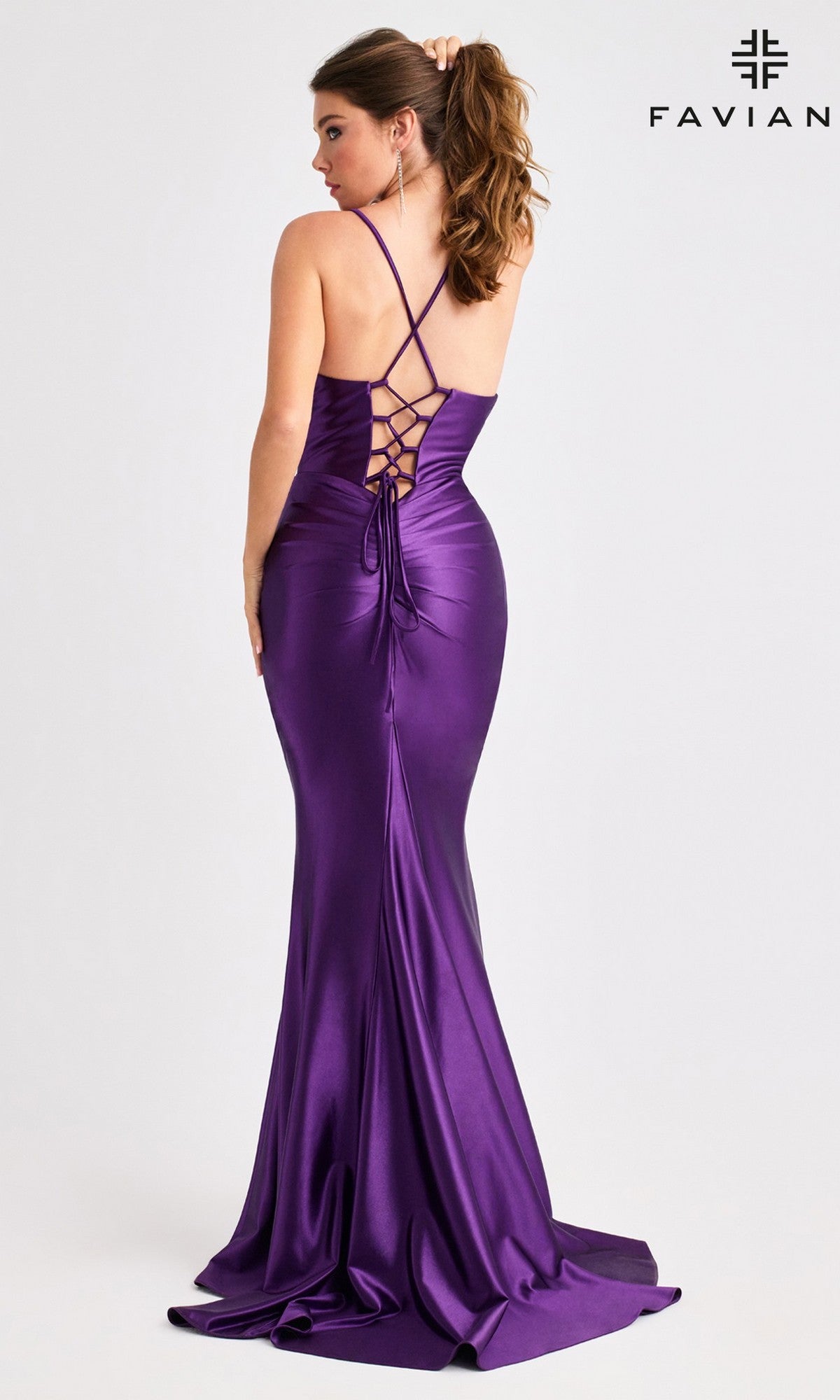 A woman models the Faviana E11423, a fitted satin long prom dress with a leg slit, lace-up open back, and crisscross straps. She stands before a plain light background with the Faviana logo in the top right corner.