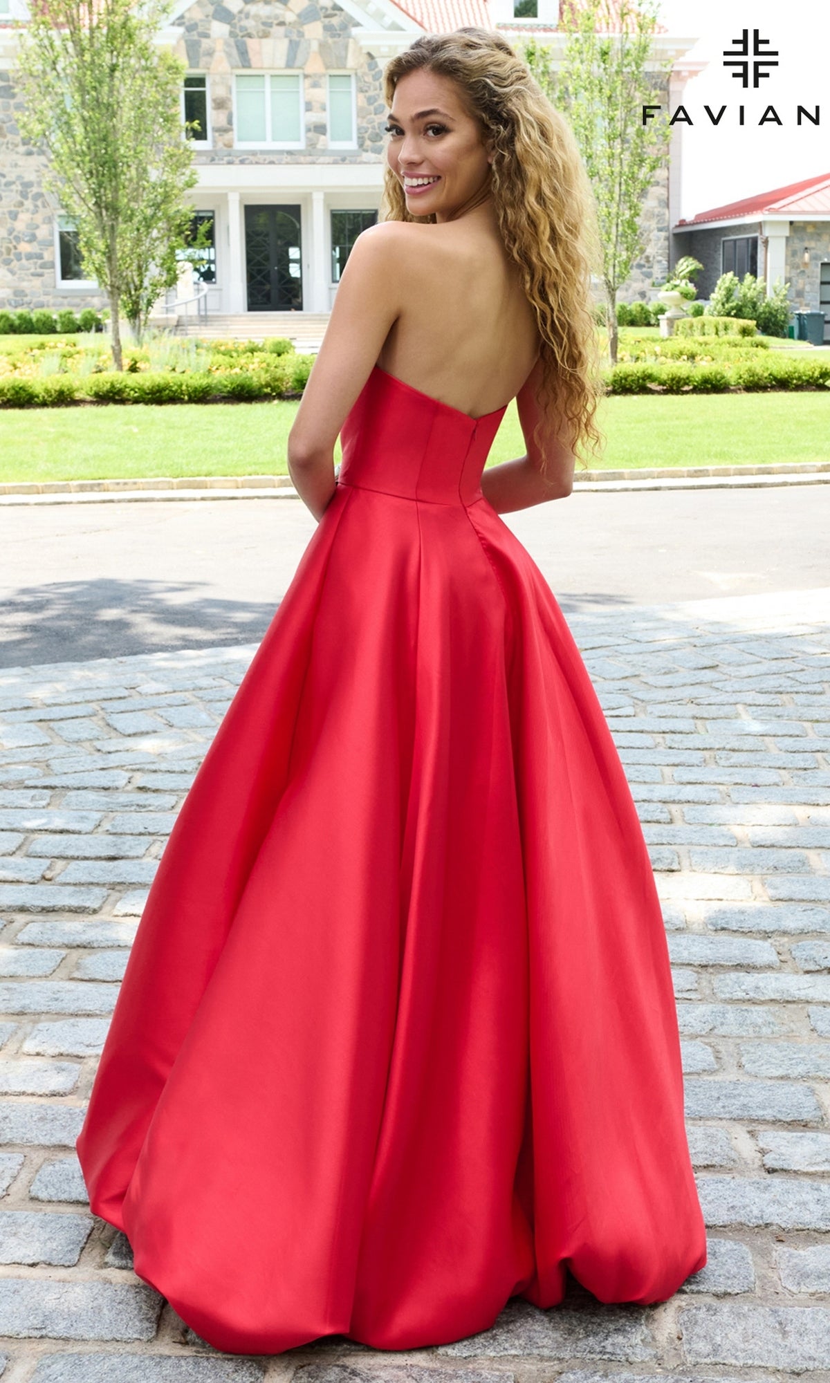 A woman on a cobblestone driveway wearing a strapless, floor-length red Faviana E11670 prom dress with a full bubble hem.