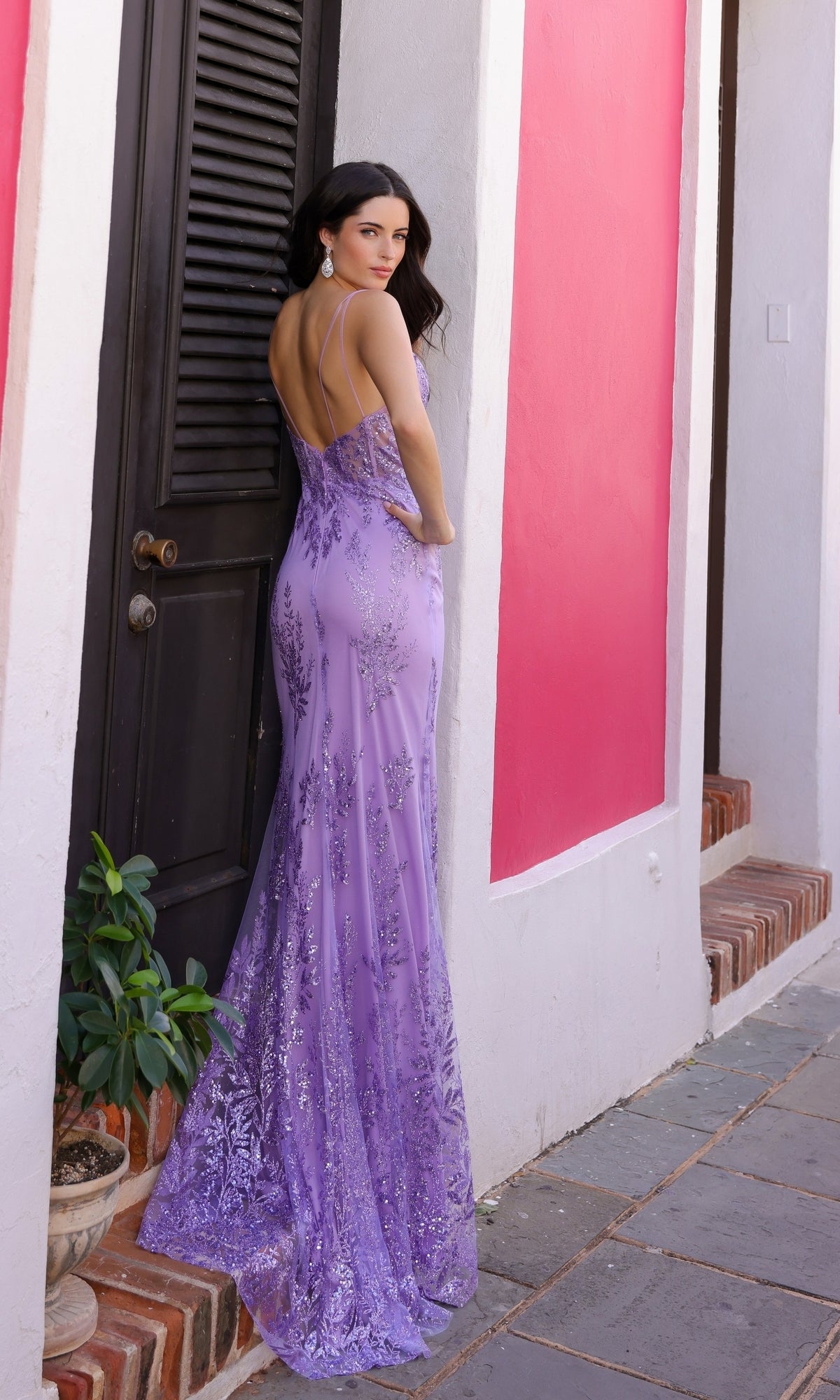 A woman wearing the Glitter-Print Long V-Neck Prom Dress E1273 stands by a black door and a pink-and-white wall, glancing over her shoulder.
